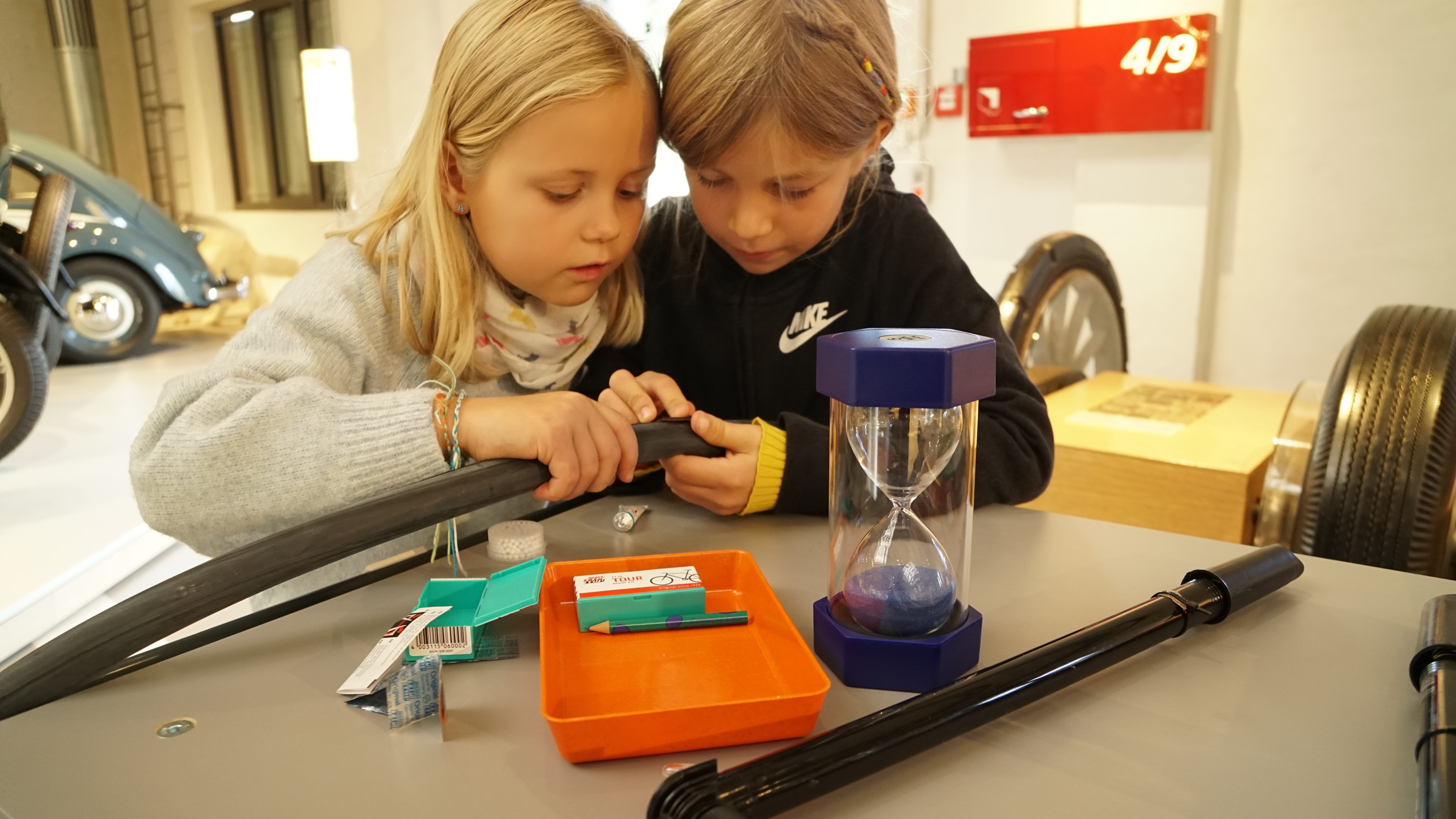 Zwei Kinder flicken in der Ausstellung einen Fahrradschlauch. Auf dem Tisch befinden sich eine Luftpumpe, eine Reifenreparatur-Set und eine Sanduhr.