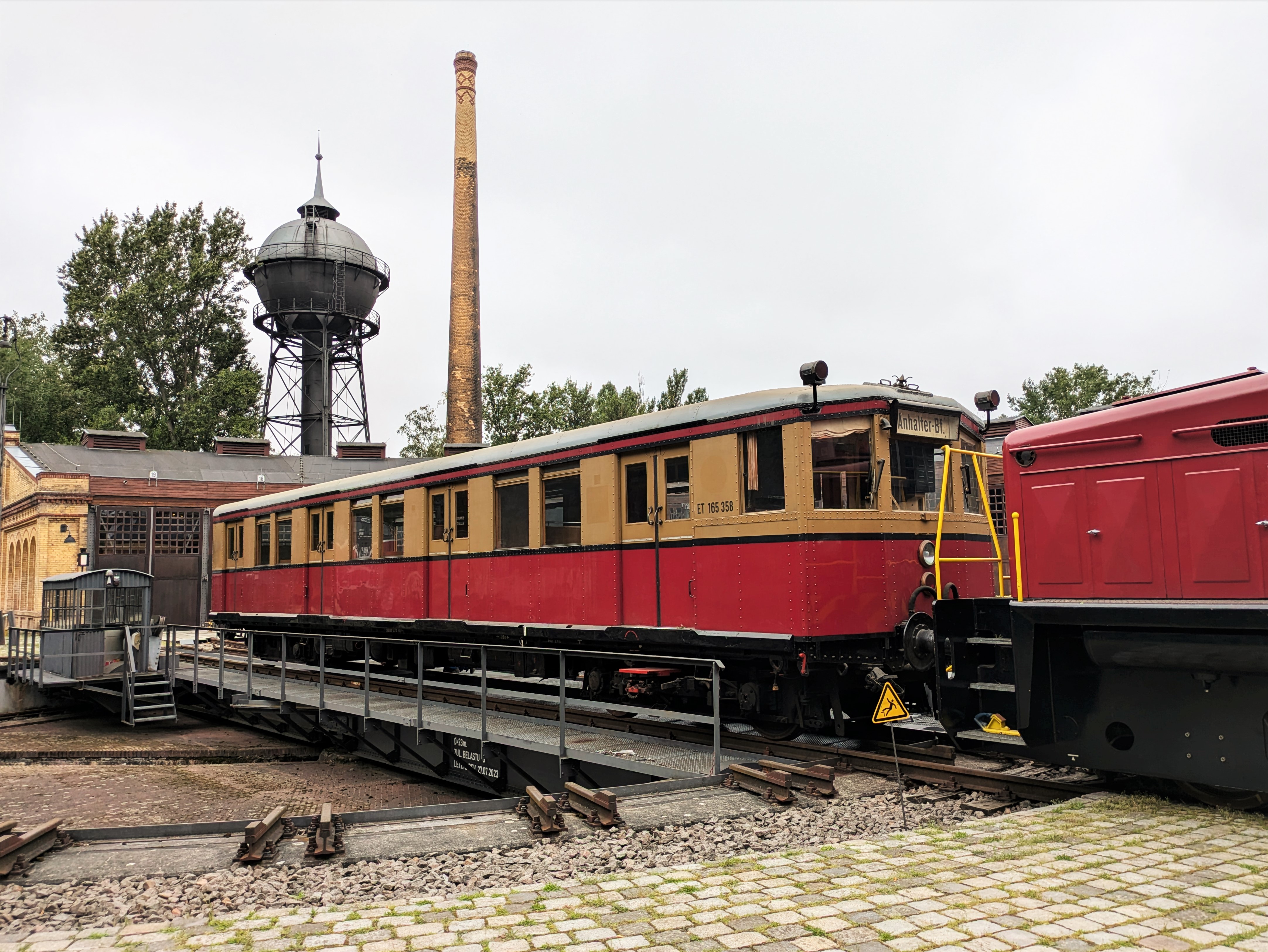 Fotos eines gelb-roten S-Bahn-Wagens auf der Drehscheibe vor einem Lokschuppen. Im Hintergrund sind ein alter Wasserturm und ein Backstein-Schornstein zu sehen.