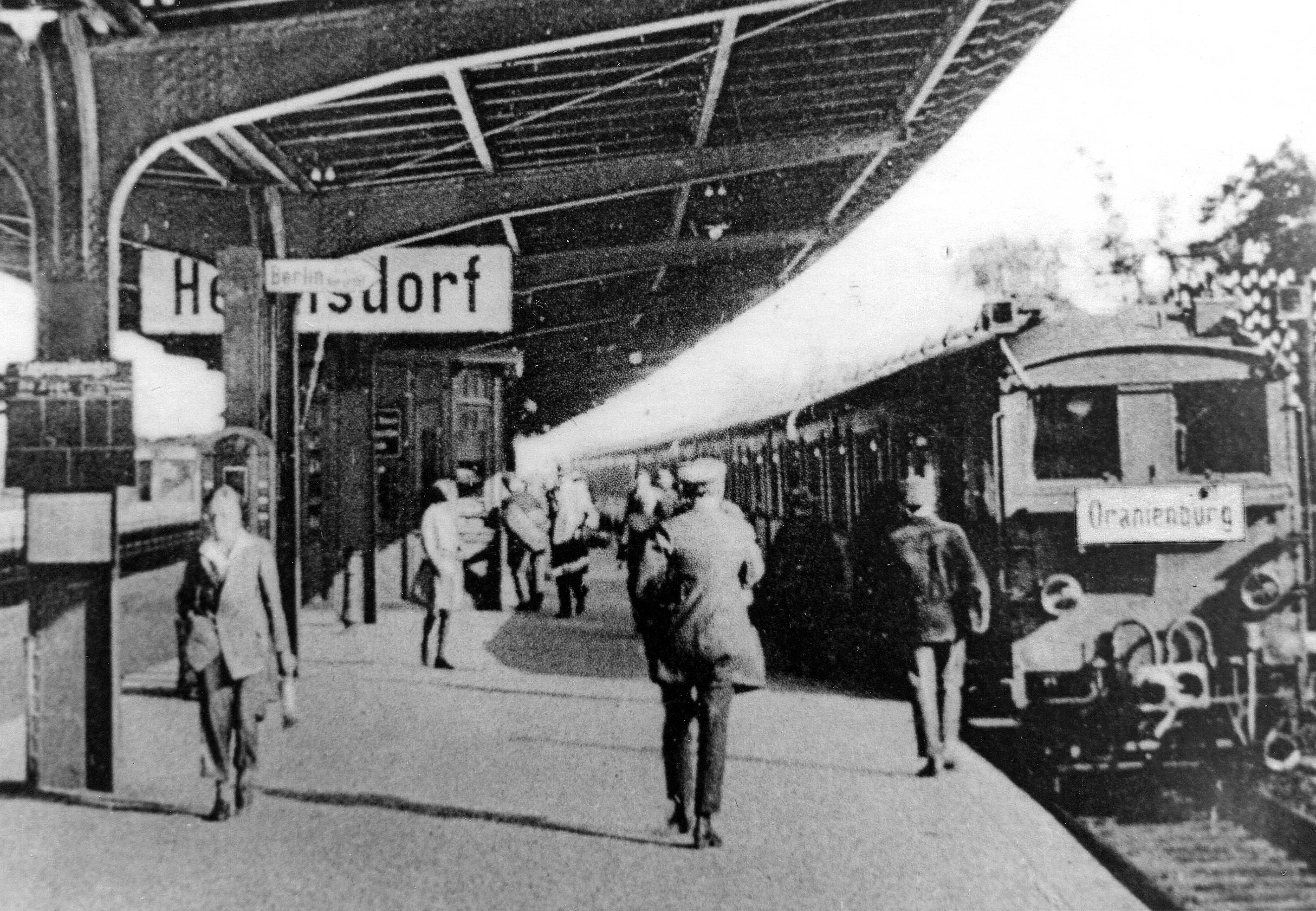 Schwarz-weiß Foto einer Szene auf einem Bahngleis um 1925. Der Schriftzug "Hermsdorf" ist zu erkennen, auf dem Gleis steht eine alte S-Bahn ein.