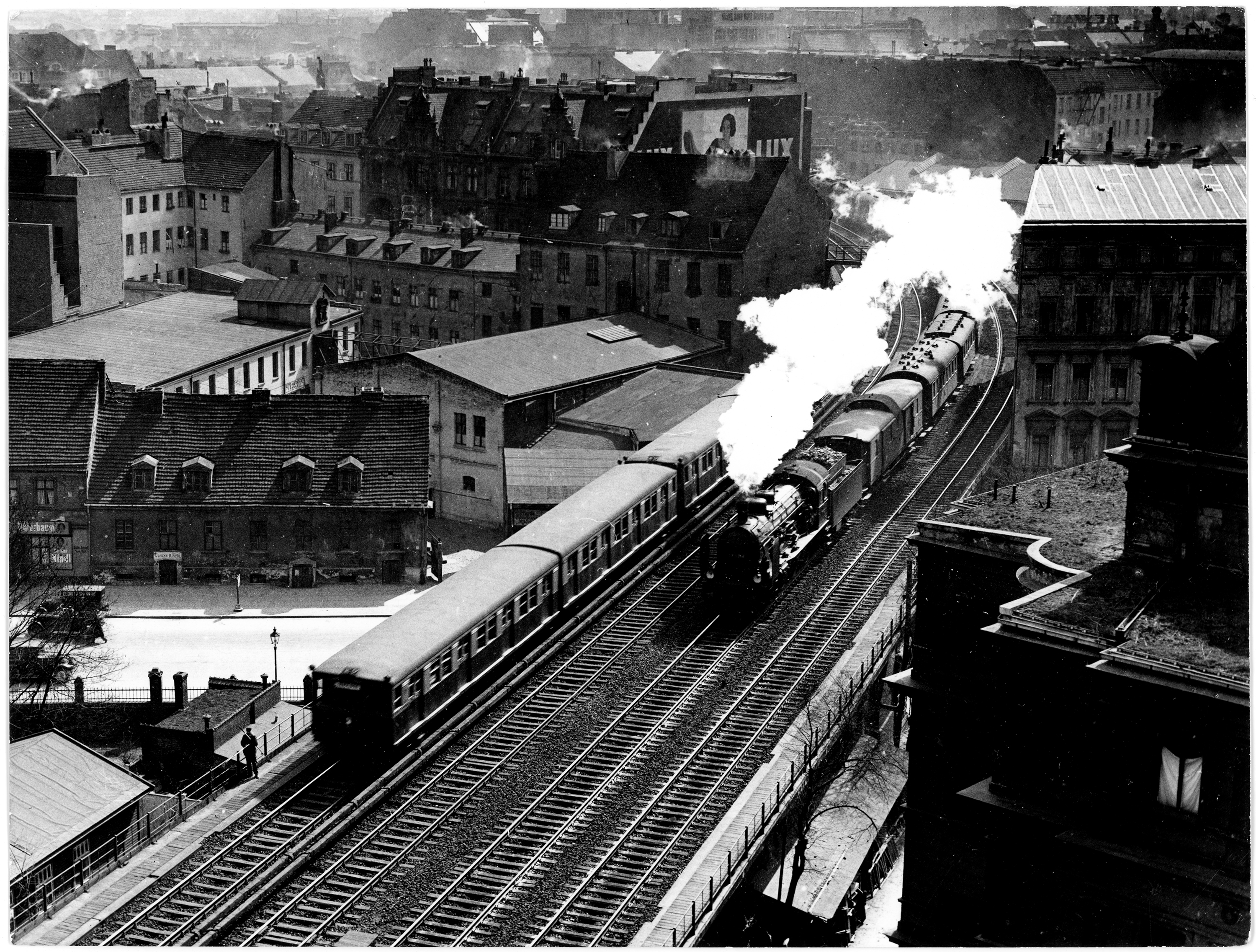 Schwarz-weiß Foto von einer Dampflokomotive und einer S-Bahn, die auf parallelen Schienen nebeneinander fahren.