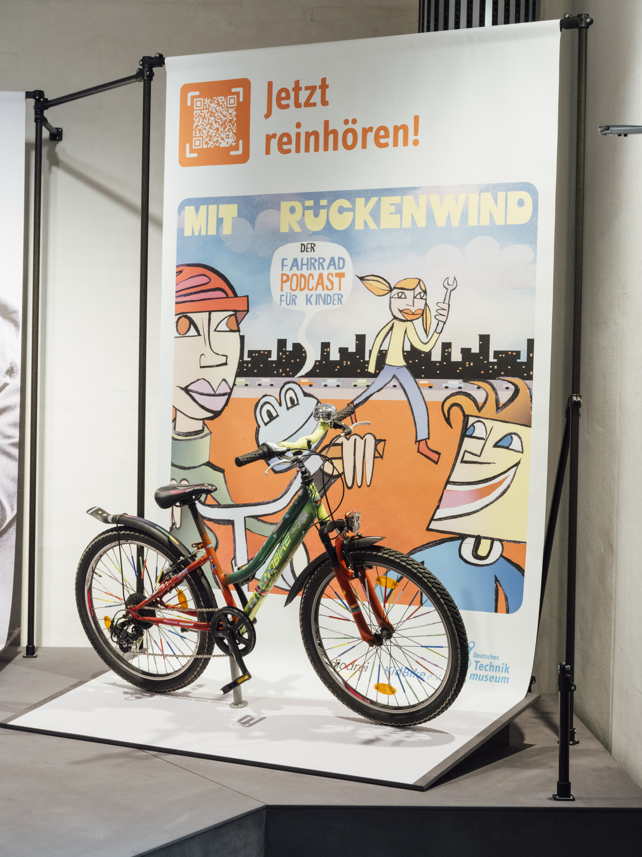 Fahrrad-Ausstellung mit Banner des Podcasts „Mit Rückenwind – Der Fahrrad Podcast für Kinder“. Buntes Fahrrad vor Plakat mit Zeichnung: Figuren, Stadtsilhouette und Slogan „Jetzt reinhören!“ mit QR-Code.