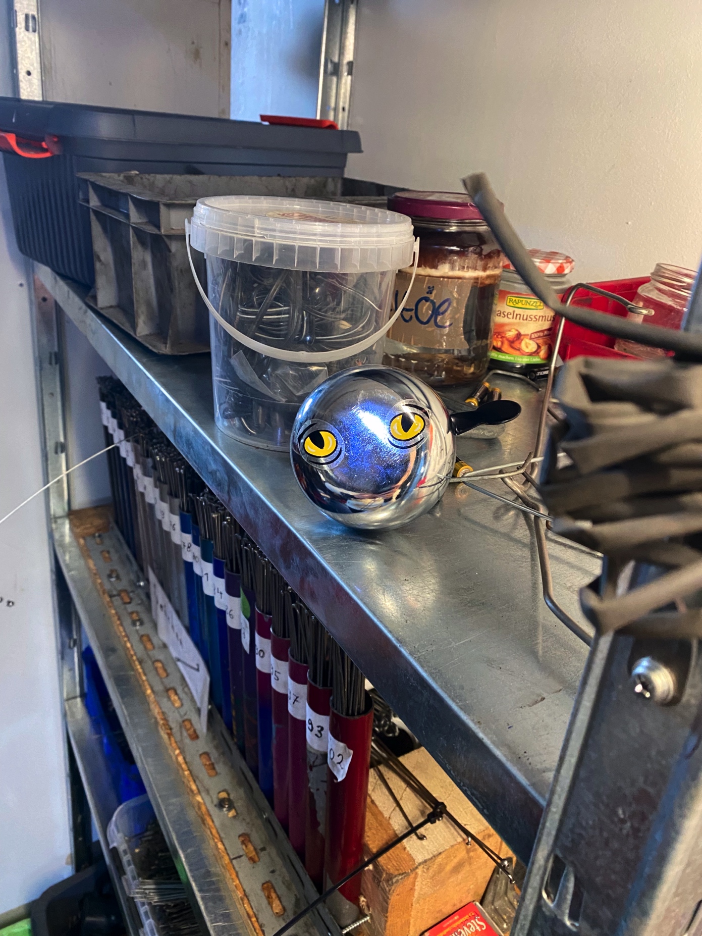 Ein Werkstattregal mit verschiedenen Gegenständen: ein silberne Fahrradklingel mit gelben Augen, Schrauben in einem Plastikbehälter, Gläser mit Aufschriften und farbige Fahrradspeichen in Halterungen.
