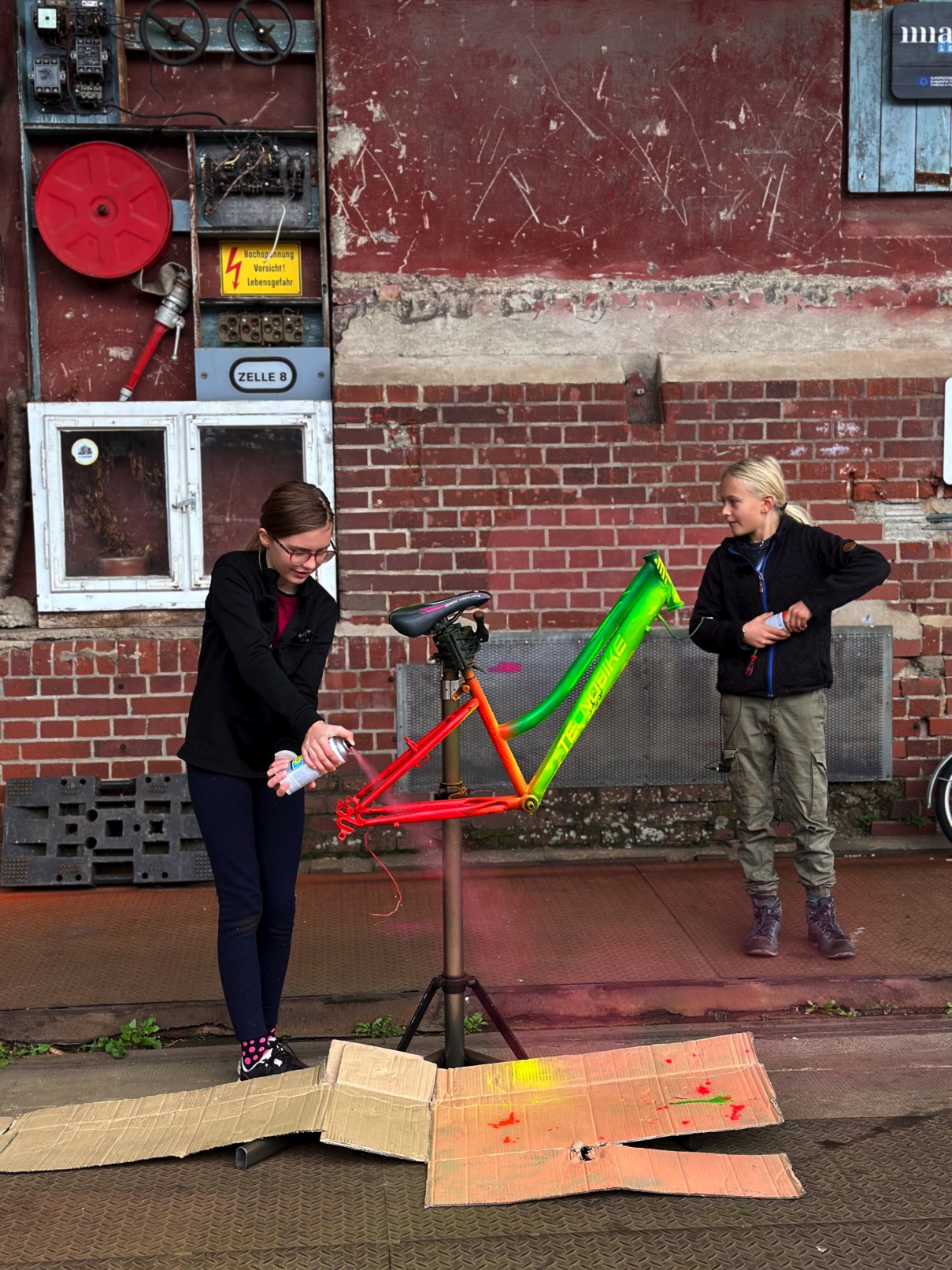 Zwei Kinder sprühen bunte Farbe auf den Rahmen eines Fahrrads, das auf einem Ständer montiert ist. Im Hintergrund ist eine Backsteinwand mit Schildern und alten Gerätschaften zu sehen.