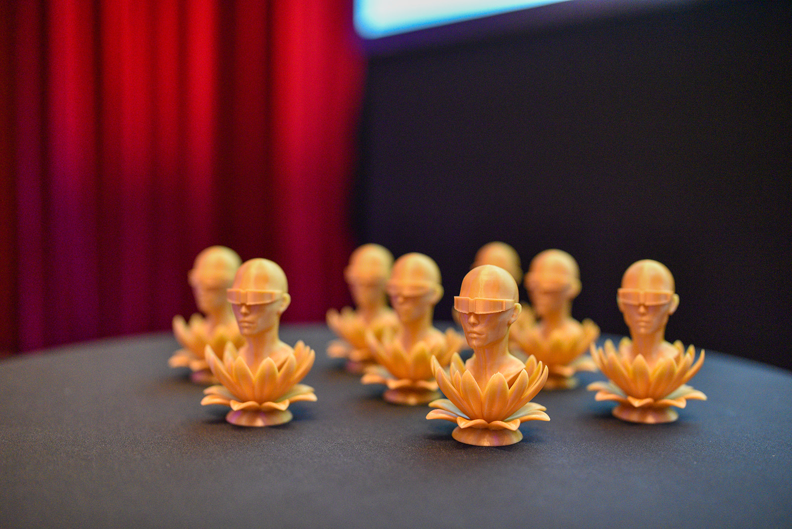 Eine Gruppe von goldfarbenen Trophäen in Form eines aus einer Lotosblüte wachsenden Kopfes mit einer VR-Brille. Die Awards stehen auf einem Tisch, im Hintergrund ist ein roter Vorhang zu sehen.