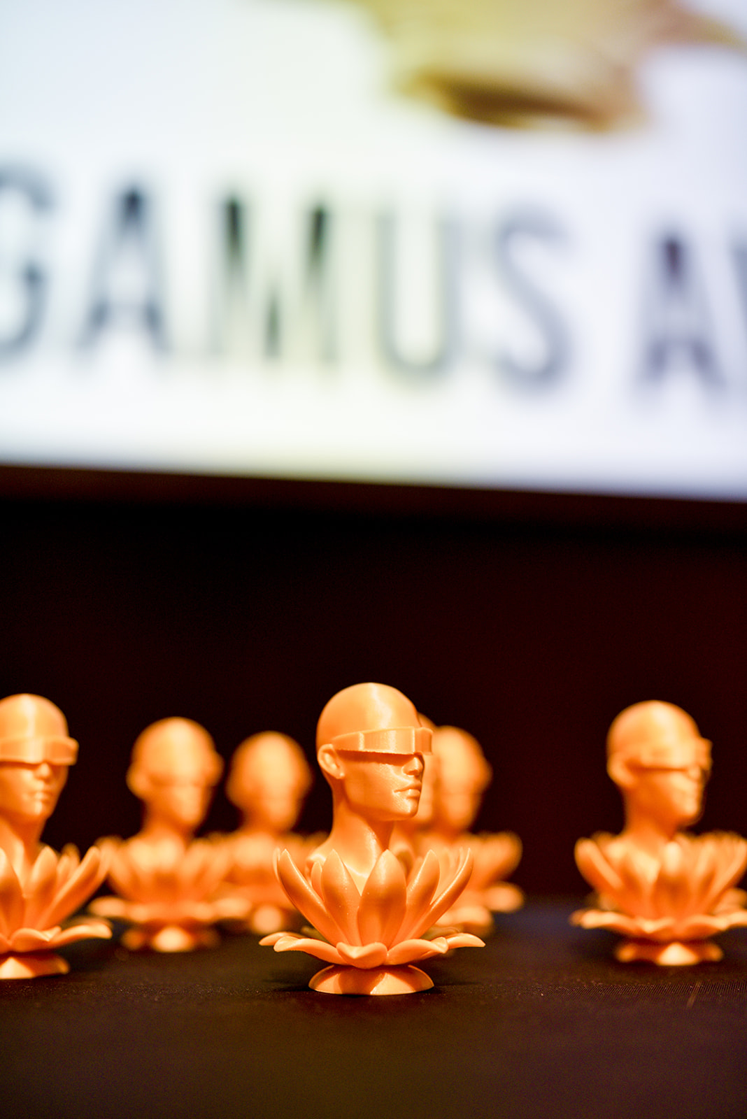 Eine Gruppe von goldfarbenen Trophäen in Form eines aus einer Lotosblüte wachsenden Kopfes mit einer VR-Brille. Die Awards stehen auf einem Tisch, im Hintergrund ist ein Teil des Schriftzuges "Digamus Award" zu lesen. zu sehen.