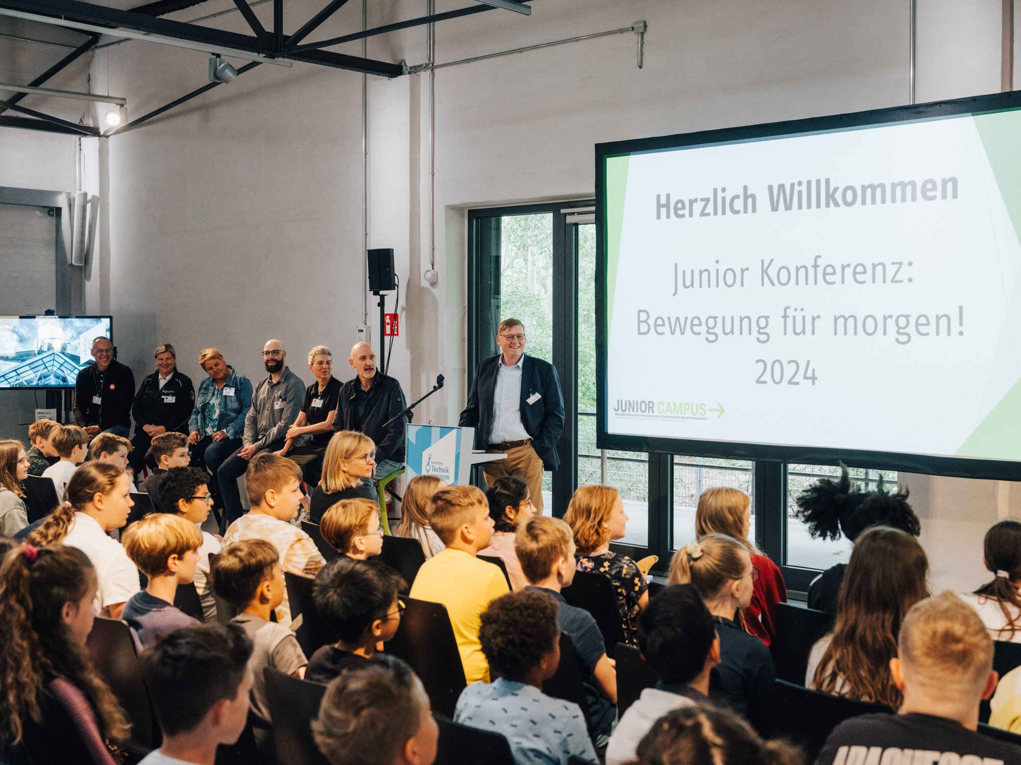 Ein Koferenz-Raum, an der Wand ein großer Bildschrim mit der Aufschrift "Hertlich willkommen, Junior Konferenz: Bewegung für morgen! 2024", die Stuhlreihen sind besetzt mit Schülerinnen und Schülern, am Stehpult steht ein Mann im Anzug und begrüßt die Kinder, links daneben sitzen sechs Erwachsene auf Stehhockern.