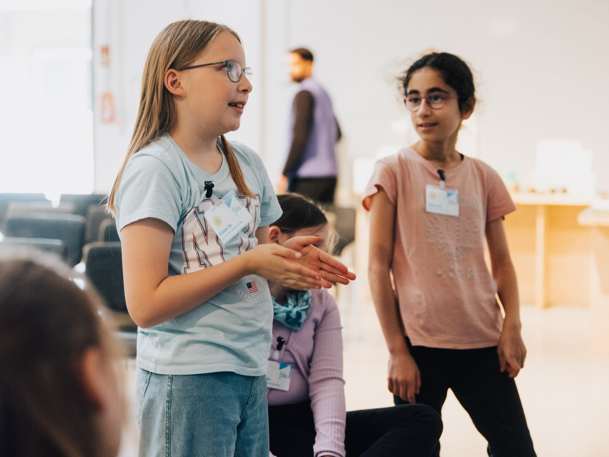Zwei Mädchen stehen in einem hellen Konferenz-Raum, das Mädchen links erklärt etwas und das andere Mädchen schaut ihm dabei aufmerksam zu. Im Hintergrund sind weitere Personen zu sehen.