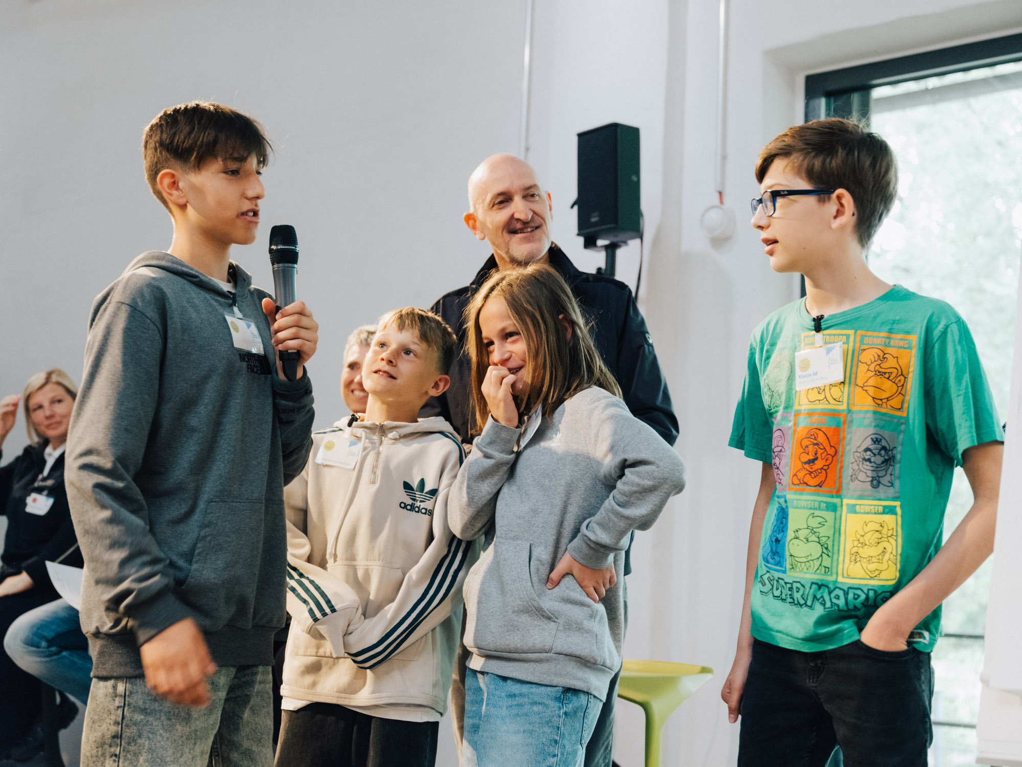 Eine Gruppe von Schülern spricht miteinander, ein Junge hält ein Mikrofon in der Hand, im Hintergrund ist ein Mann zu sehen, der den Kindern zuhört.