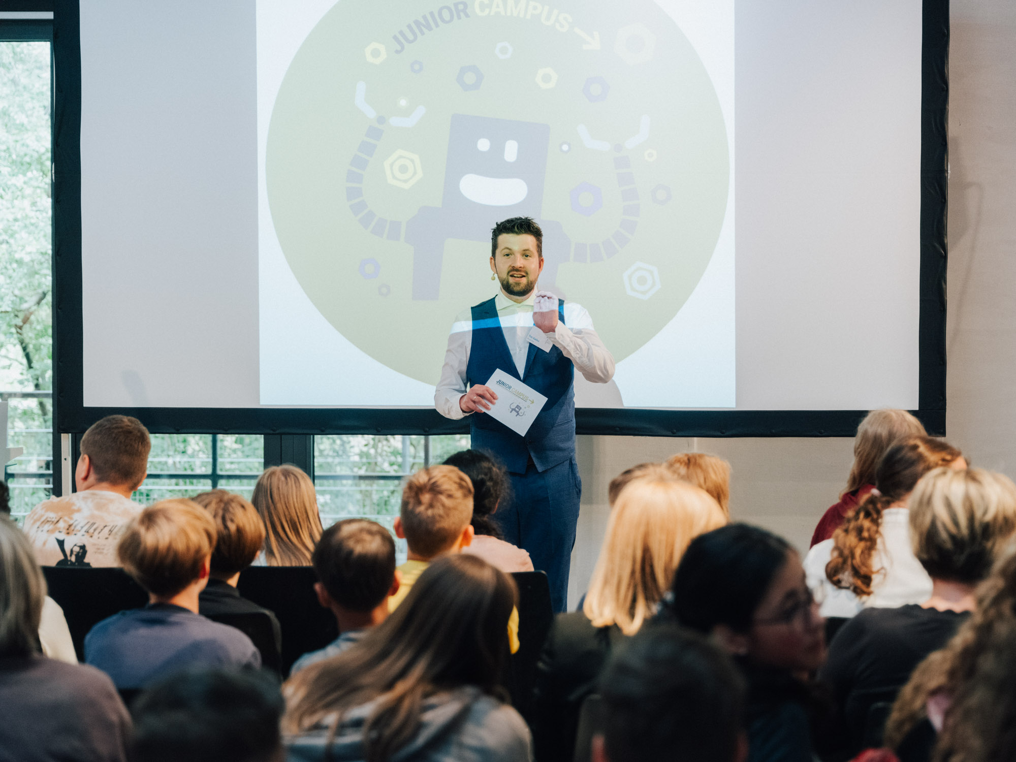 Ein offenbar gut gelaunter Mann im Anzug steht vor einer Leinwand auf der ein lustiger Roboter und die Aufschrift "Junior Campus" zu sehen sind. Vor ihm sitzen Schülerinnen und Schüler in Stuhlreihen und hören ihm gebannt zu.
