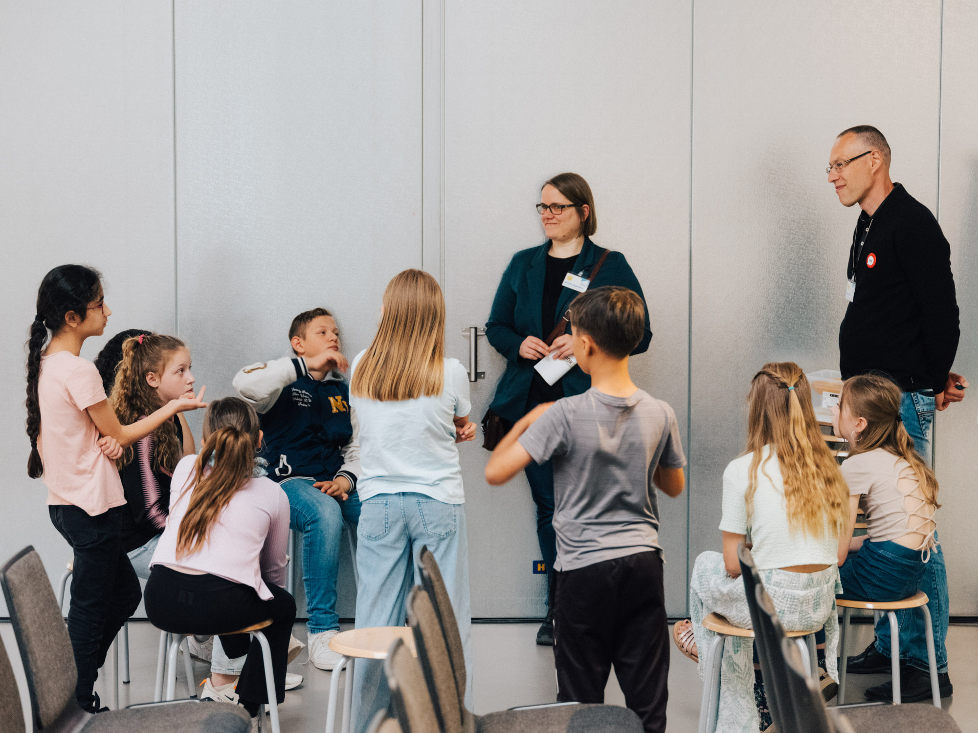 Eine Gruppe von Schülerinnen und Schülern steht mt zwei Erwachsenen in einem raum. Sie unterhalten sich, ein Mädchen links erklärt etwas, die anderen Personen hören aufmerksam zu.