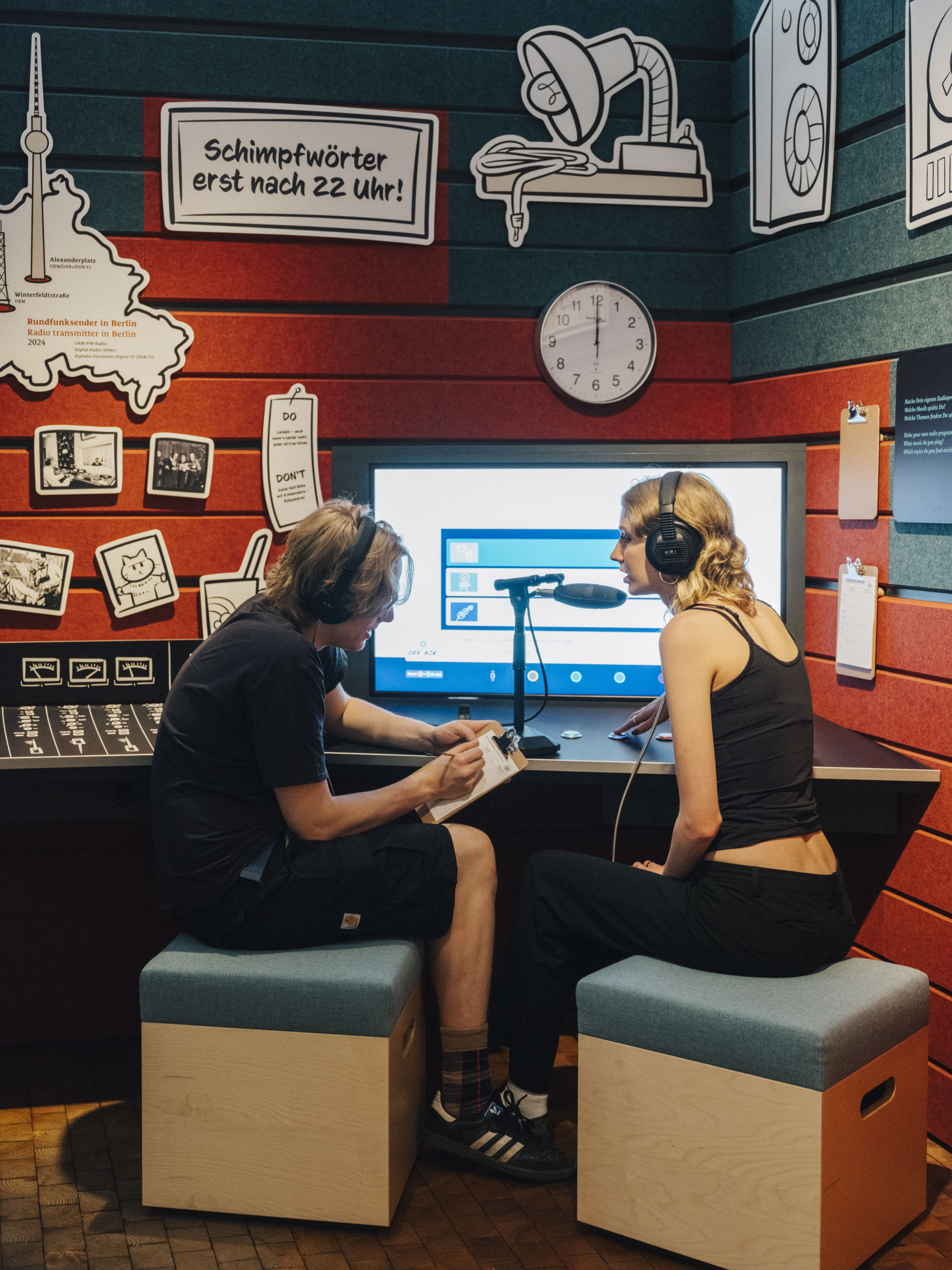 Das Innere des gläsernen Studios im Ausstellungsbereich "Radio Gleisdreieck". Im Studio sind zwei Personen mit Kopfhörern und Mikrofon dabei, eine Sendung zu produzieren.