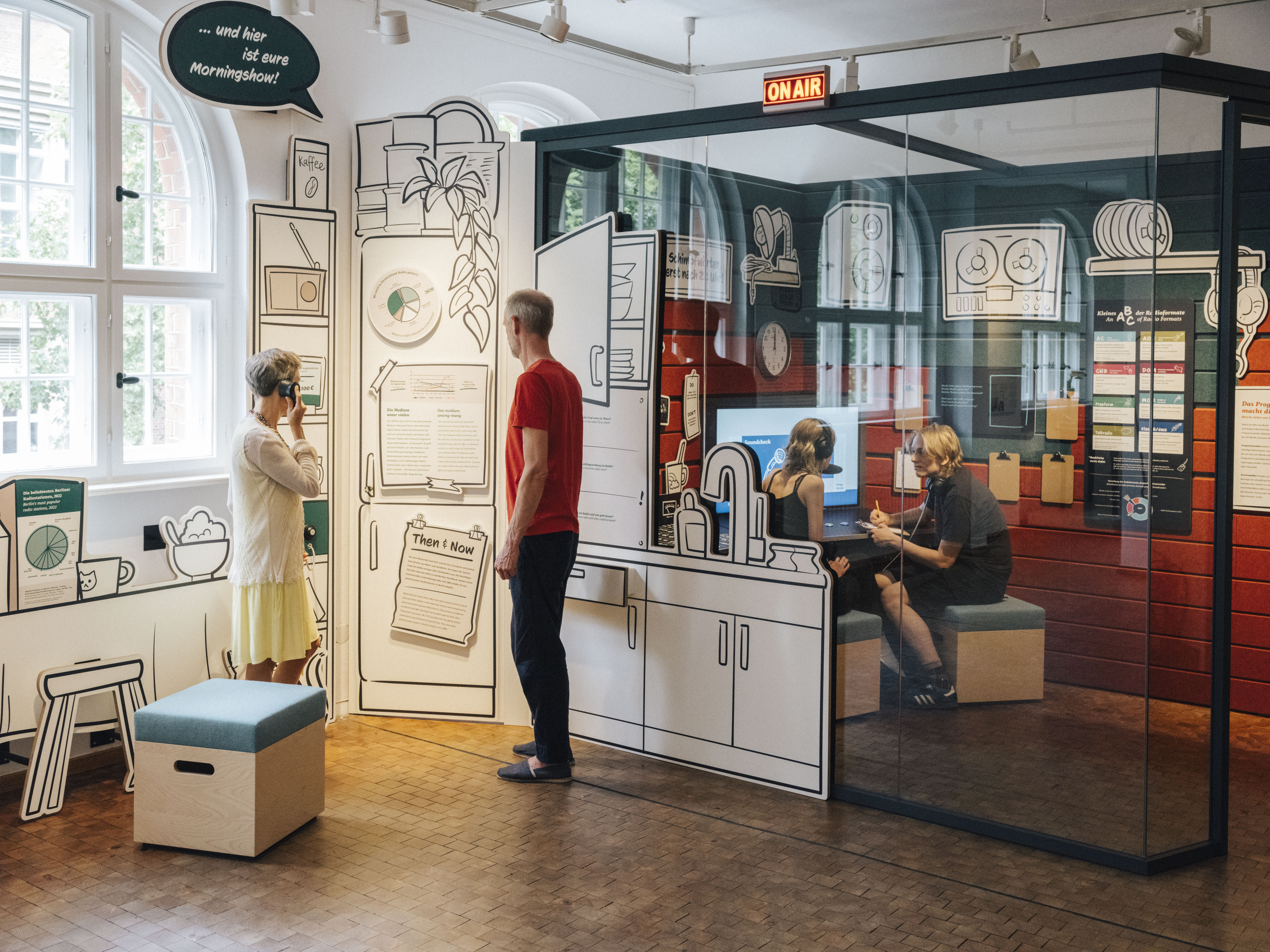 Blick in das gläserne Studio im Ausstellungsbereich "Radio Gleisdreieck". Im Studio sind zwei Personen zu erkennen. Vor dem Studio stehen zwei Menschen und hören etwas über Kopfhörer. Über dem Eingang zum Studio leuchtet der Schriftzug "On Air".