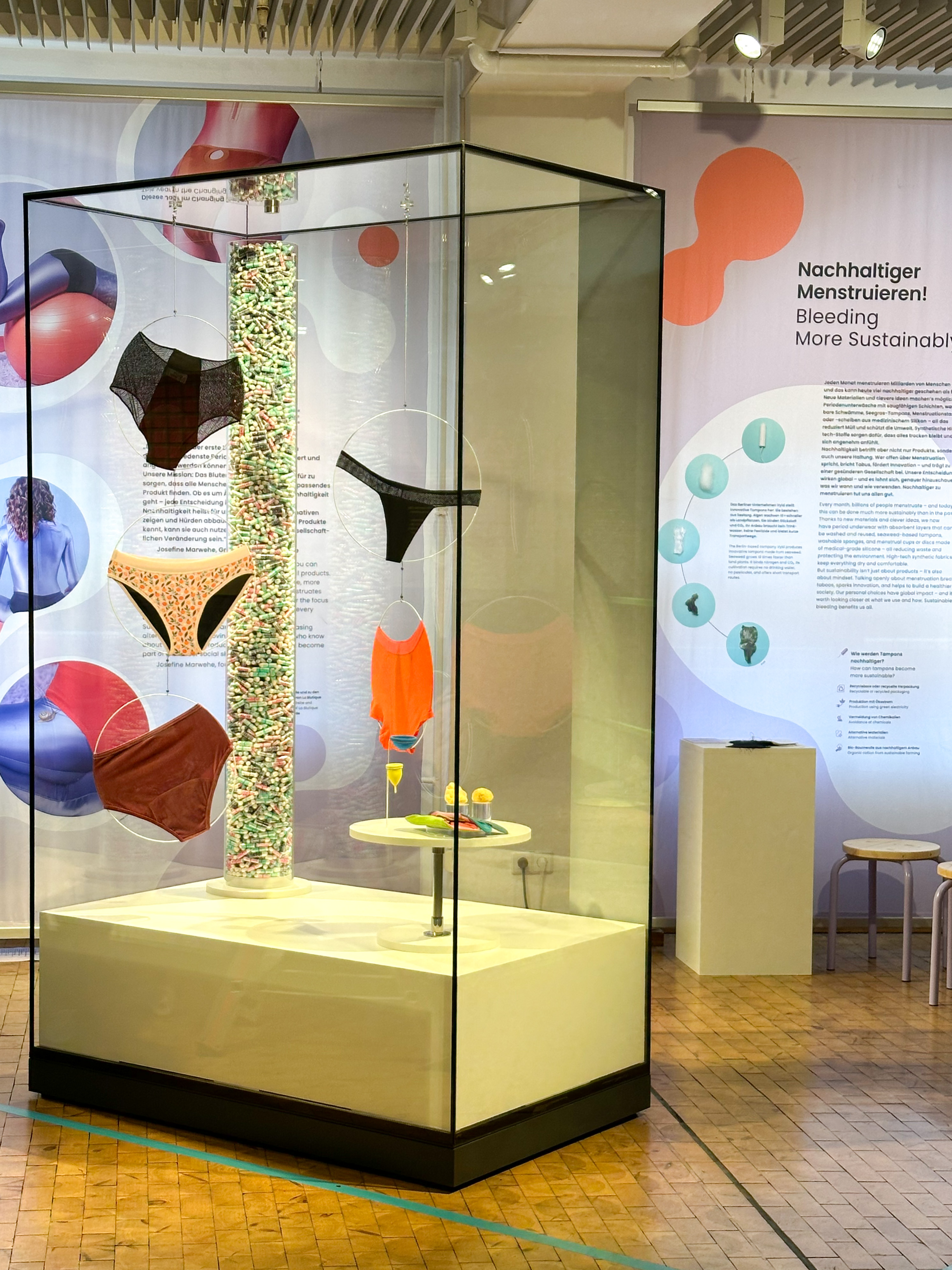 Blick auf die Vitrine im Ausstellungsbereich "Changing Room": Zu sehen sind verschiedene Modelle von Perioden-Unterwäsche, Textil-Binden, Menstruations-Cups und Tampons. Im Hintergrund ist der Schriftzug "Nachhaltiger menstruieren!" zu lesen.