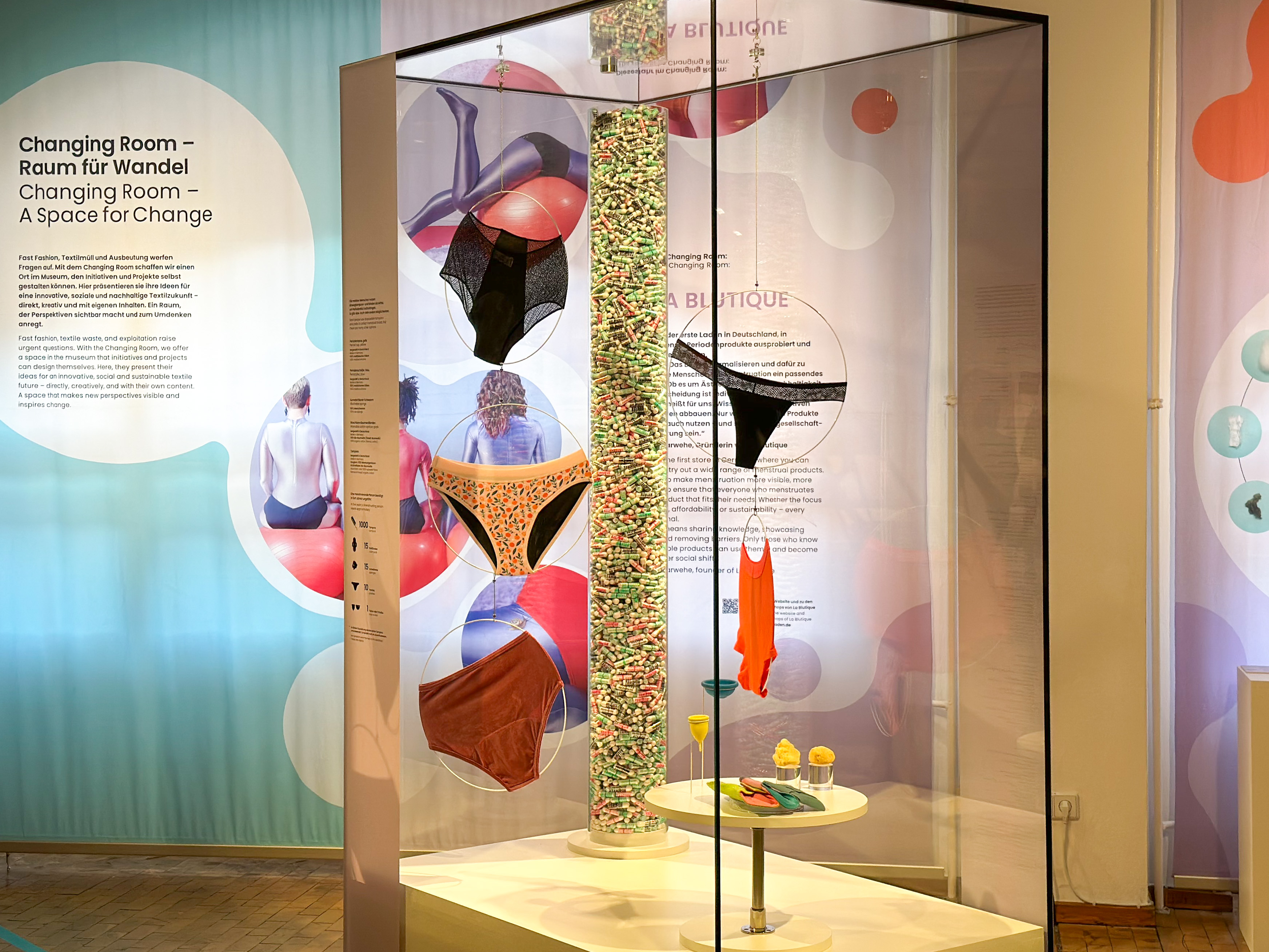 Blick auf die Vitrine im Ausstellungsbereich "Changing Room": Zu sehen sind verschiedene Modelle von Perioden-Unterwäsche, Textil-Binden, Menstruations-Cups und Tampons. Im Hintergrund ist der Schriftzug "Changing Room" zu lesen.