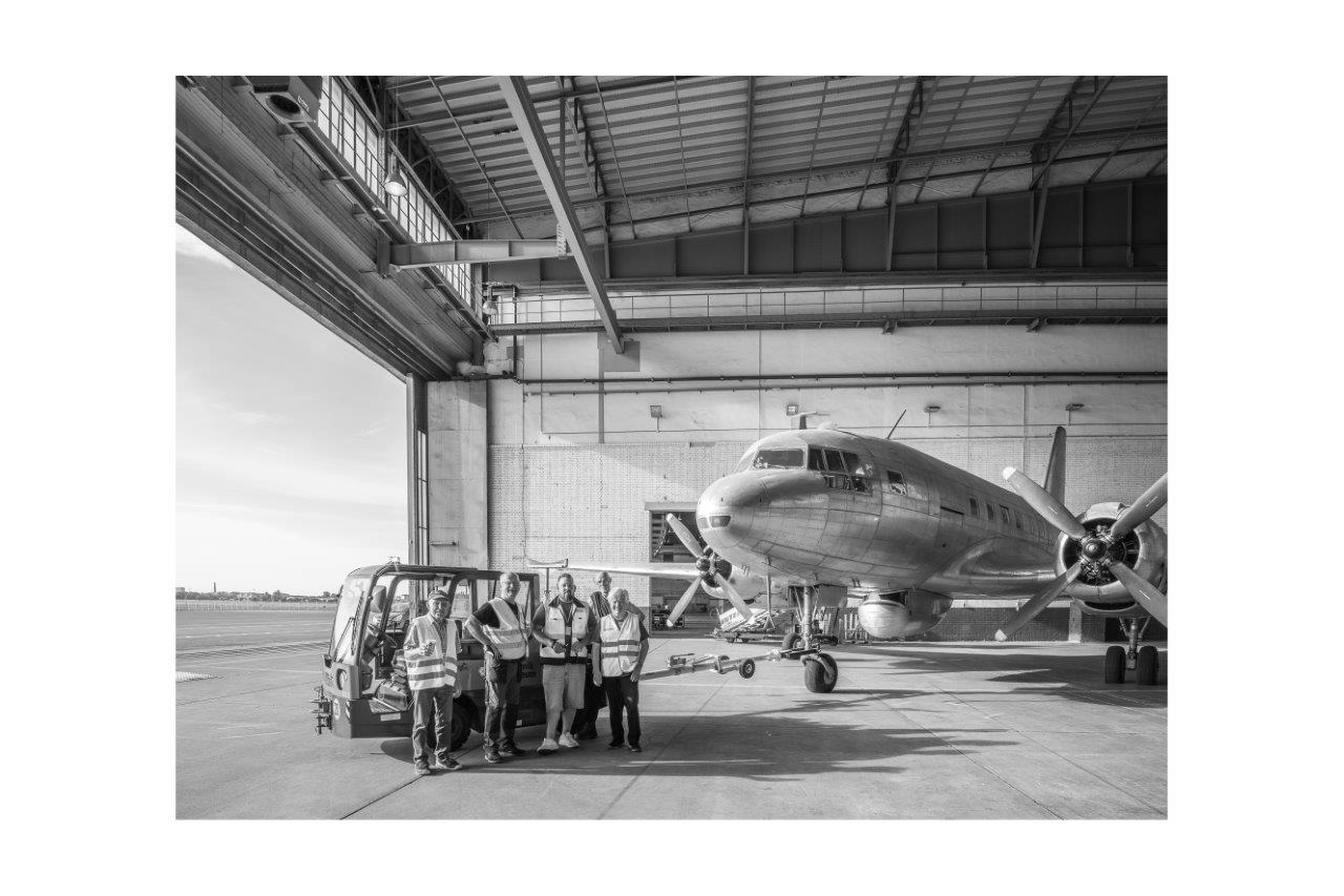 Schwarzweiss-Foto: Ein Gruppe älterer Herren steht vor einer Iljuschin in einem Hangar.