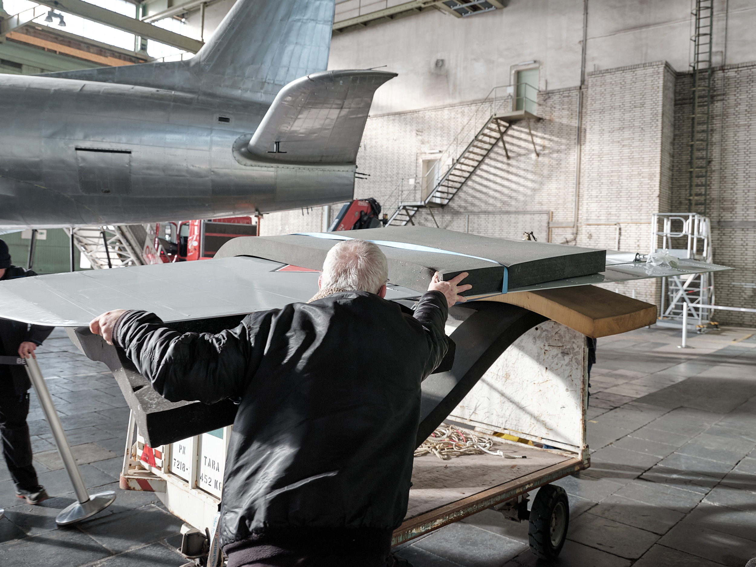 Ein älterer Herr hält eine Tragfläche eines Flugzeugs in den Händen, im Hintergrund ist der Rumpf des Flugzeugs zu sehen.