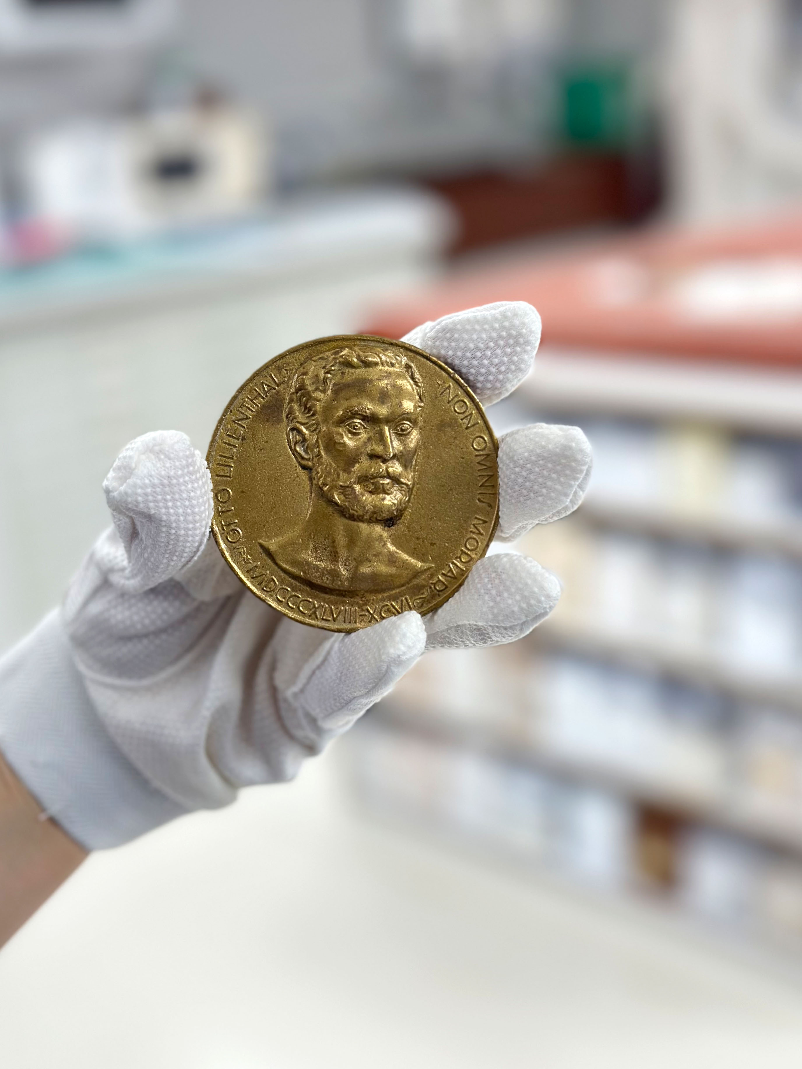 Eine weiß behandschuhte Hand hält eine messingfarbene Medaille. Darauf ist das Gesicht eines bärtigen Mannes abgebildet, am Rand stehen die Worte „Otto Lilienthal“, mehrere römische Jahreszahlen und die Worte „Non omnis moriar“.