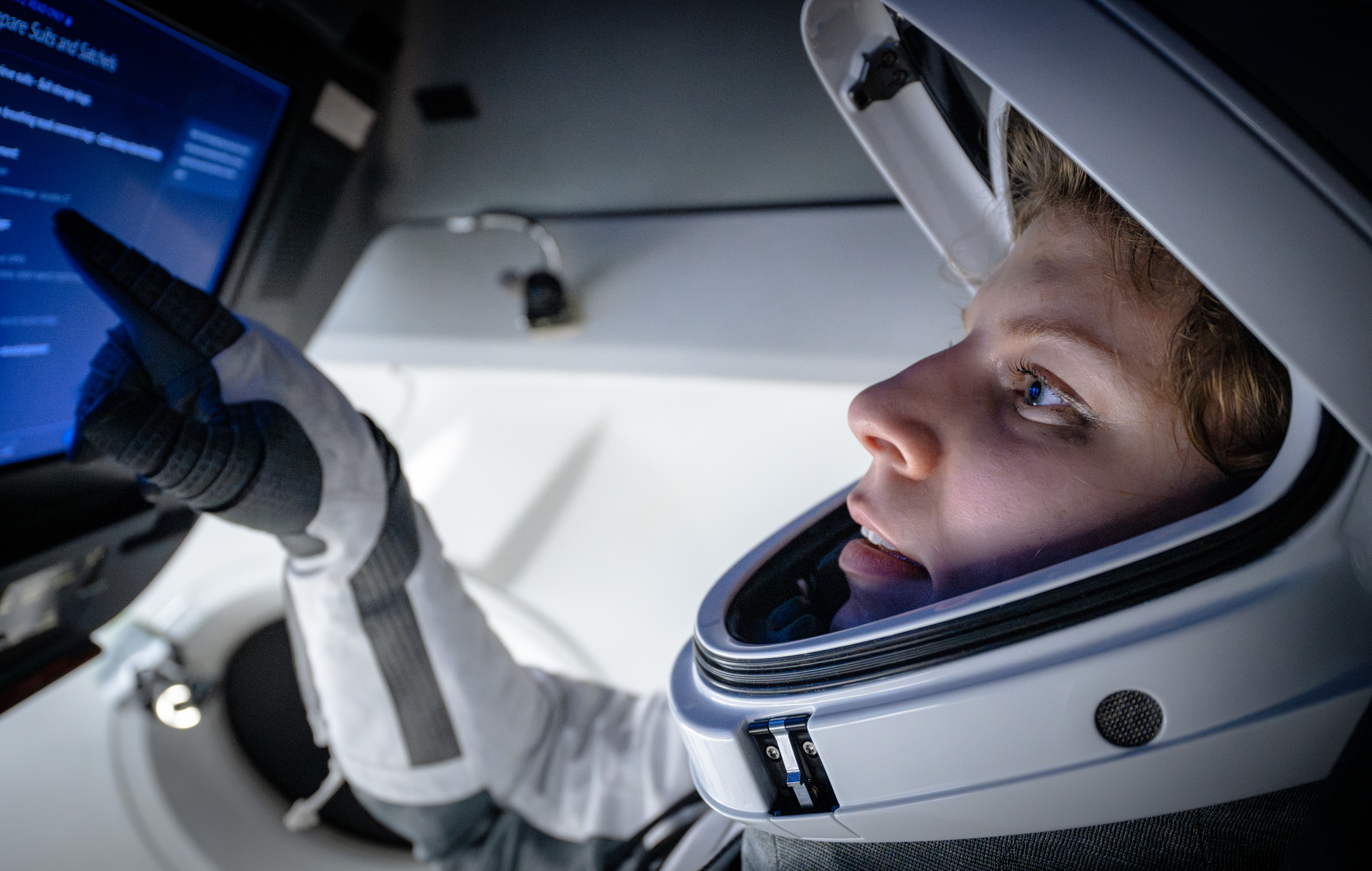 Close up des Gesichts einer jungen Person mit weißem Astronauten-Helm auf dem Kopf. Mit einem schwarz-weißen Handschuh zeigt die Person auf einen blauen Bildschirm.