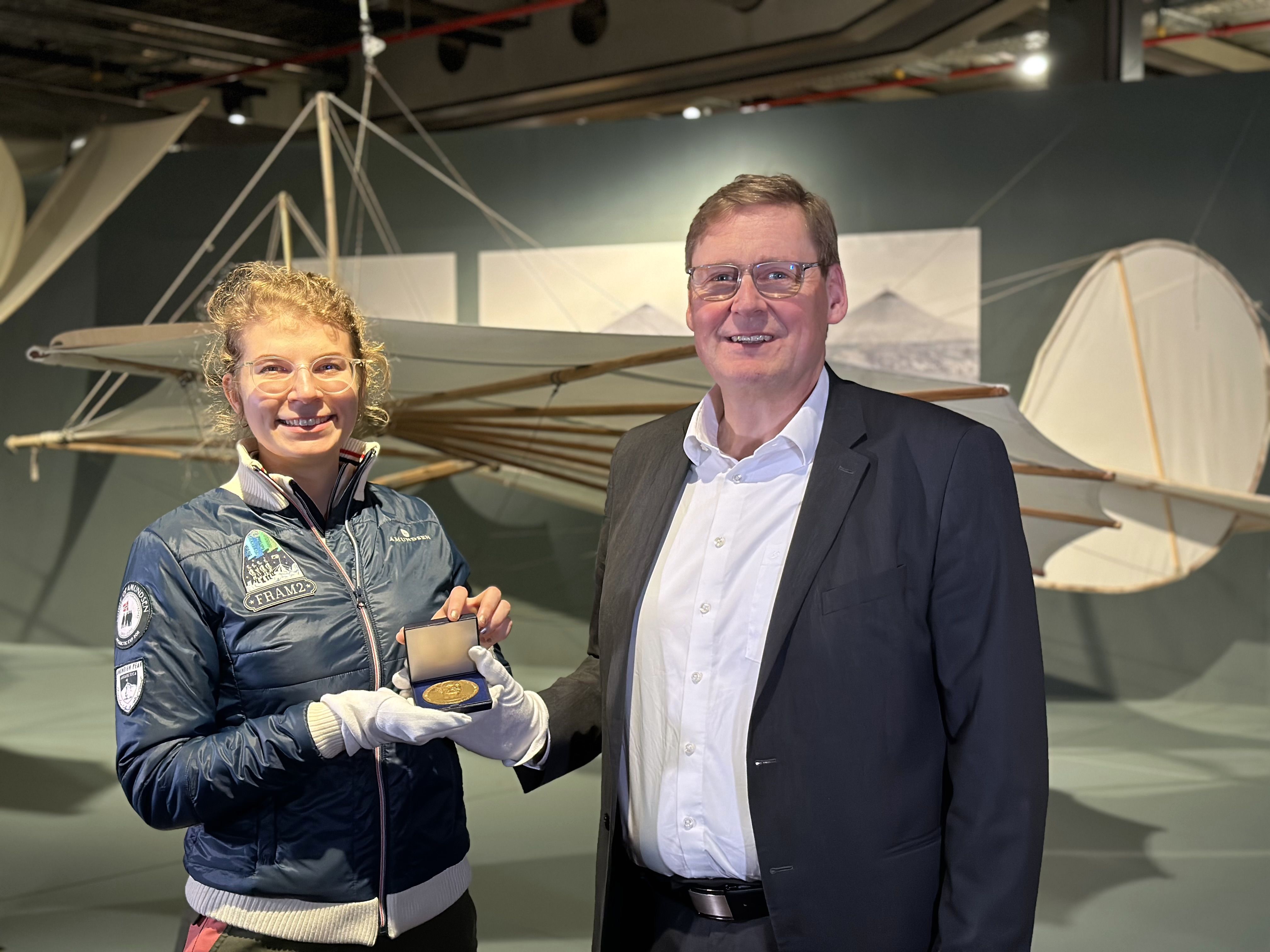 Eine blonde Frau und ein blonder Mann stehen nebeneinander in einem Ausstellungsraum. In den Händen halten sie ein aufgeklapptes Kästchen, in dem eine Medaille liegt. Im Hintergrund ist ein altertümlicher Flugapparat zu sehen.