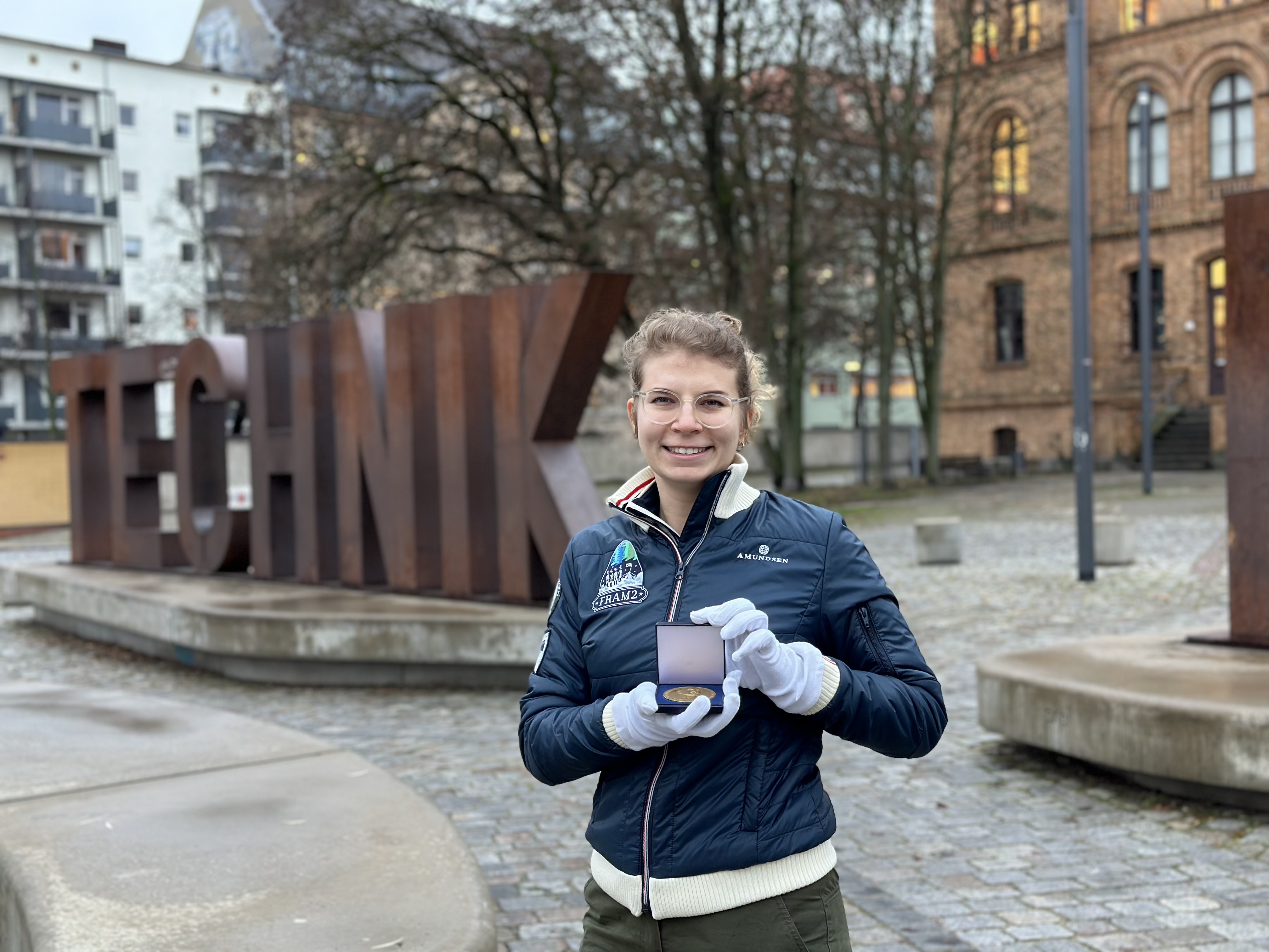 Eine junge blonde Frau in einer blauen Jacke steht lächelnd im Freien vor großen metallenen Buchstaben, die das Wort „Technik“ bilden. In der Hand hält sie ein kleines aufgeklapptes Kästchen. Darin liegt eine messingfarbene Medaille.