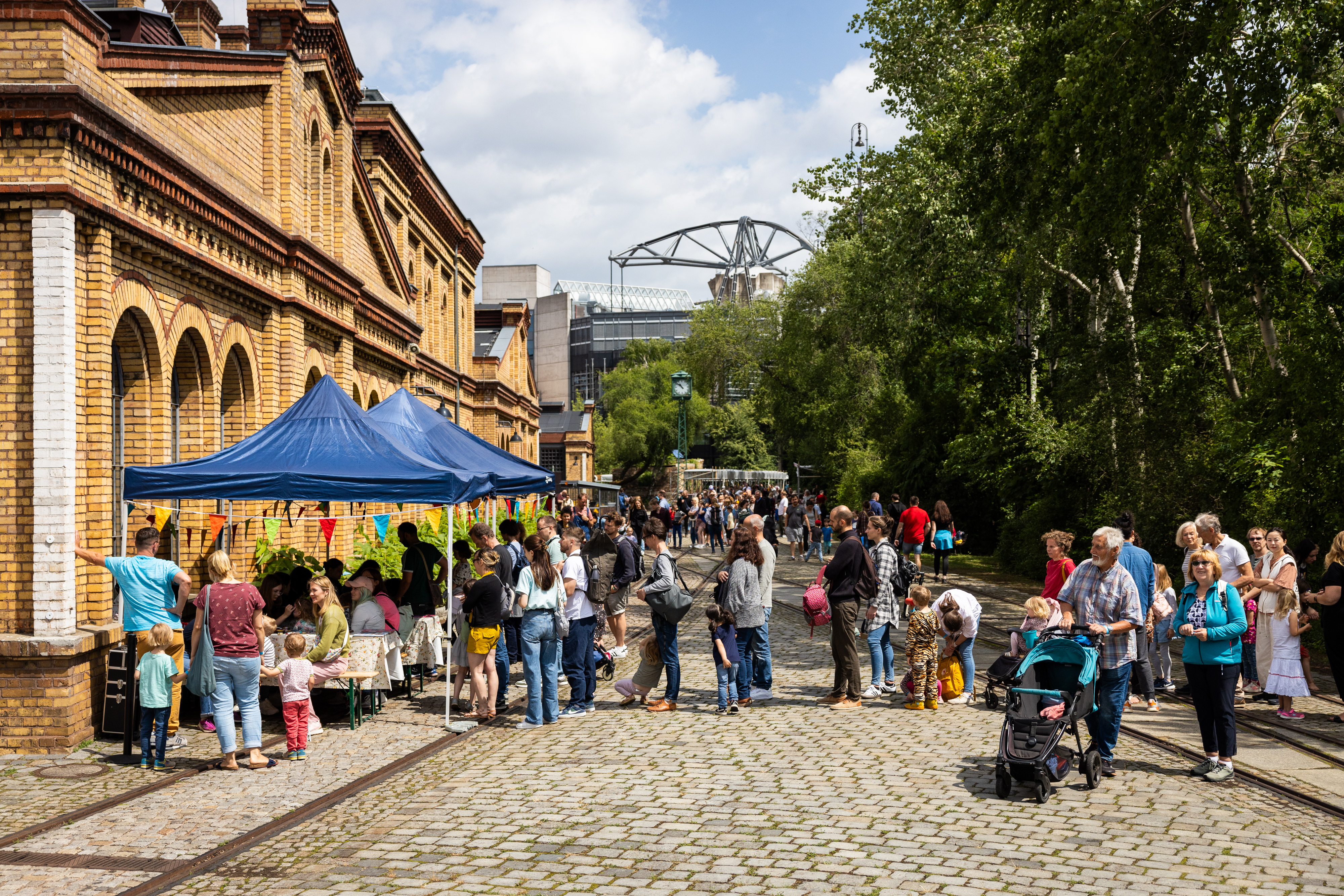 Viele Menschen in einem Park, links steht ein altes Backsteingebäude, davor ein Stand mit Sonnenschirm.