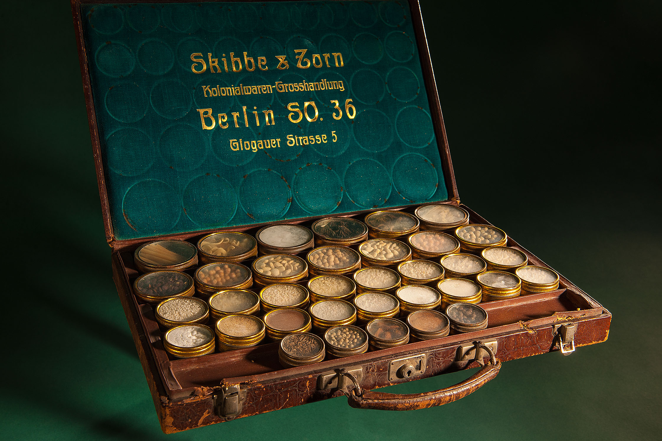 A leather case containing various small containers holding samples of colonial products such as cocoa. The inside of the case lid bears the company name Skibbe & Zorn.