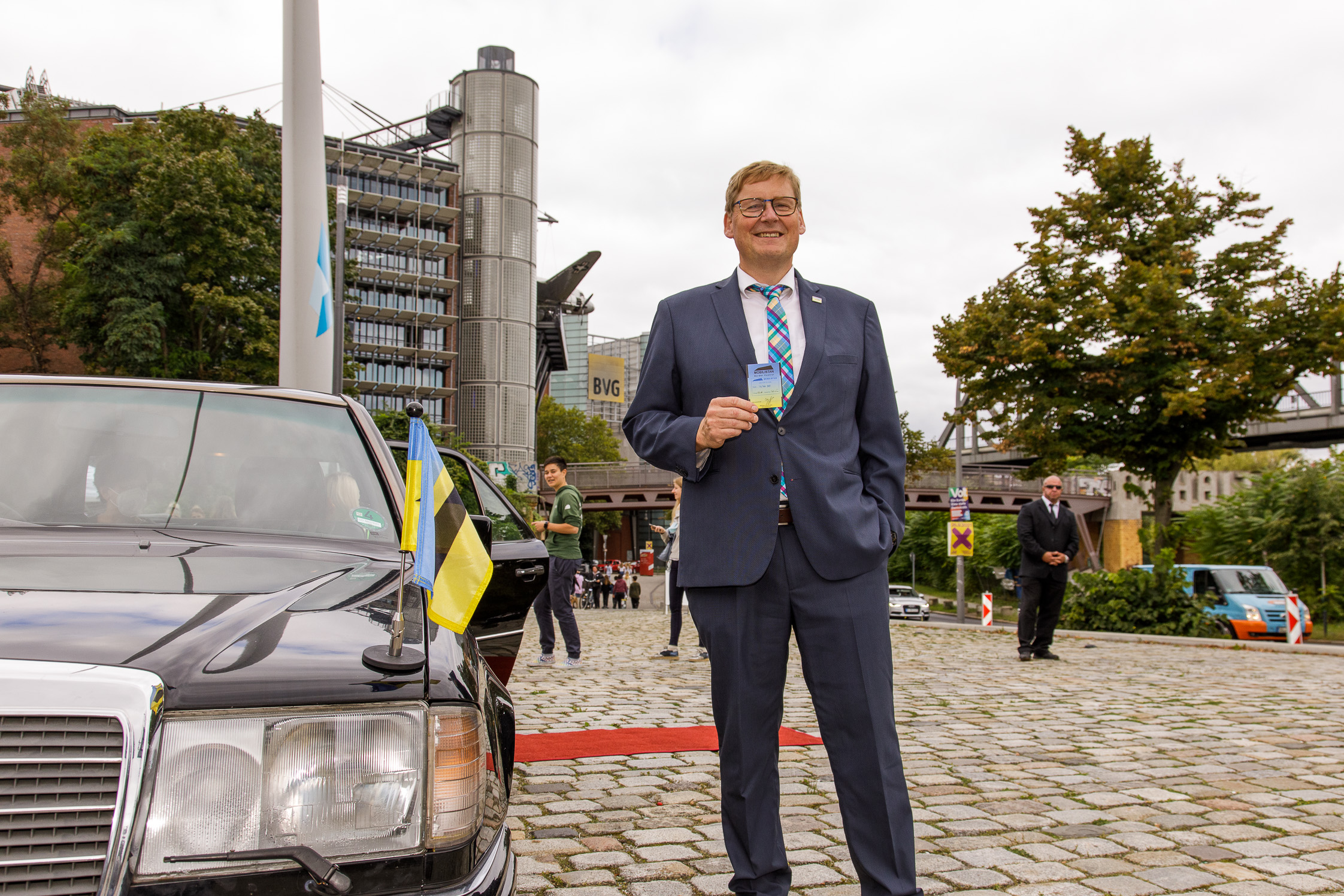 Ein Mann steht neben einer schwarzen Limousine und hält eine blaugelbe Karte in die Kamera.