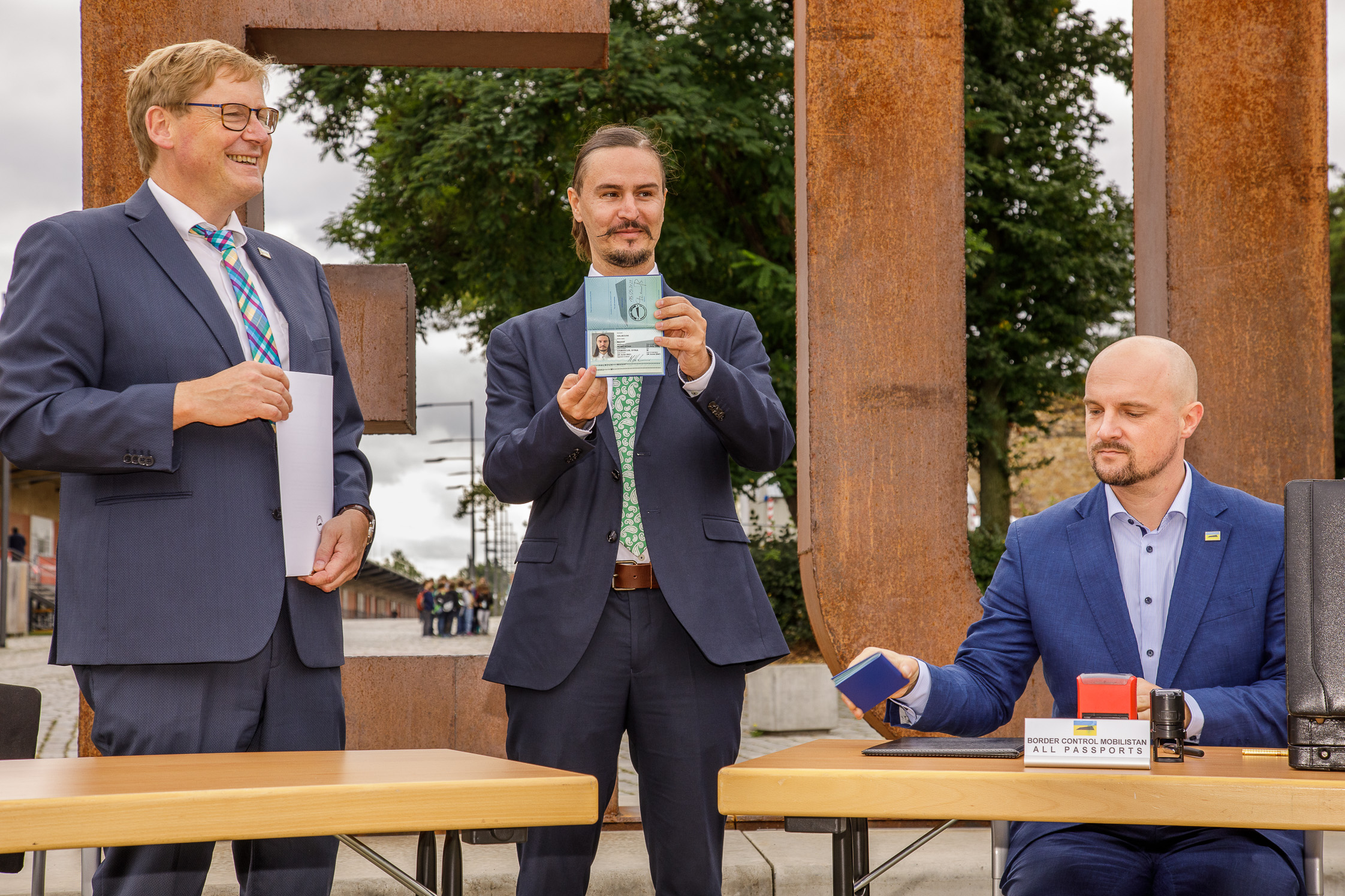 Drei Männer in Anzügen, der erste hält ein Dokument, der zweite hält den Reisepass von Mobilistan in die Höhe und der dritte sitzt am Tisch und stempelt Visa.