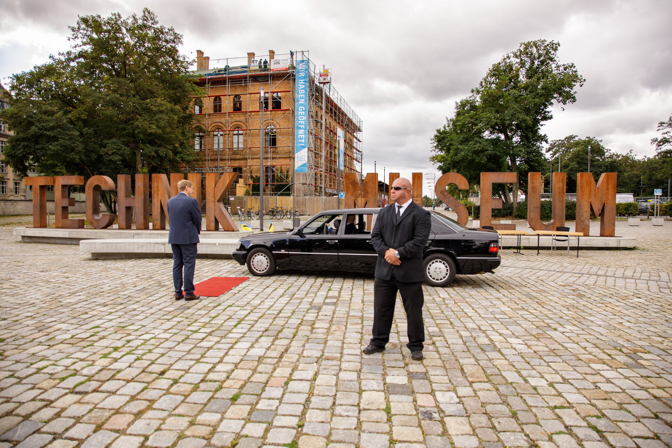 Schwarze Limousine auf dem Vorplatz, Schriftzug "Technik Museum" im Hintergrund, zwei Männer in schwarzen Anzügen.