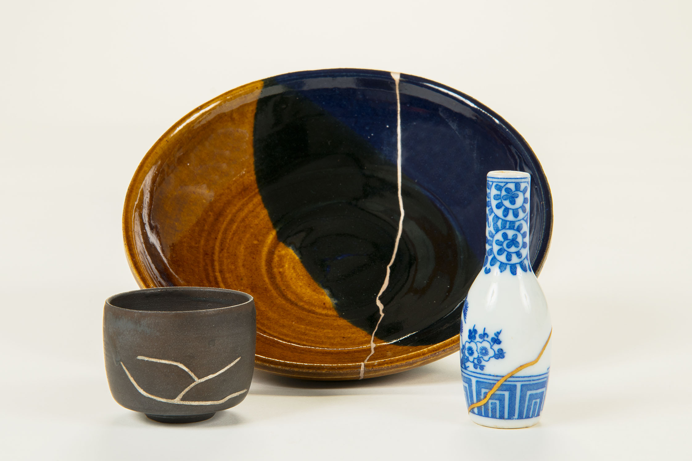 The photo shows three ceramic pieces that were obviously broken but have been repaired with gold-colored putty. In the center is a brown-and-black plate with a golden crack running down the middle; to the right is a blue-and-white vase with a crack patched with gold; and to the left is a brown bowl with several cracks filled with gold-colored putty. 