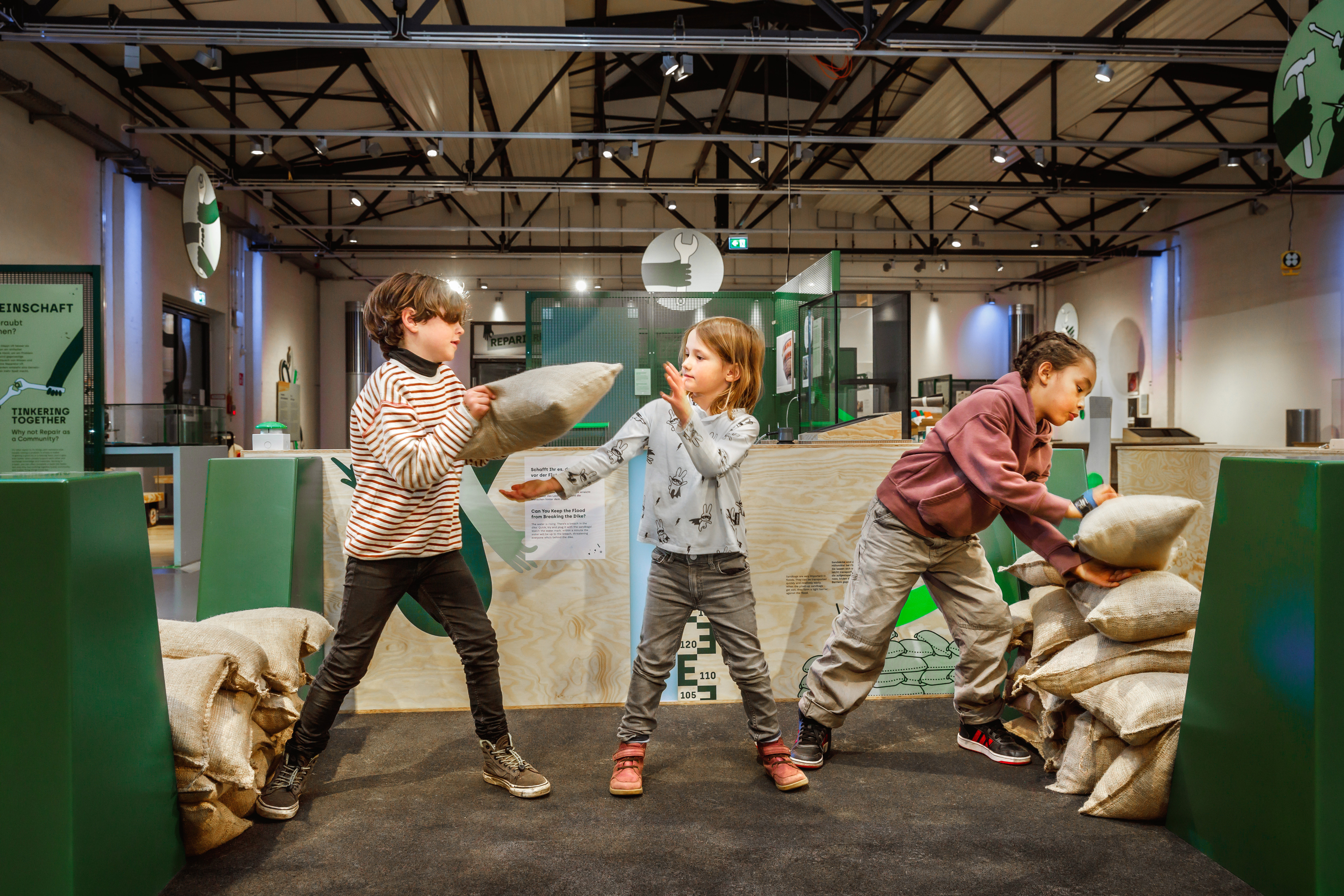 Three children are repairing a toy dike made of sandbags in an exhibition hall. They form a chain and pass the bags to one another to patch a hole in the dike.