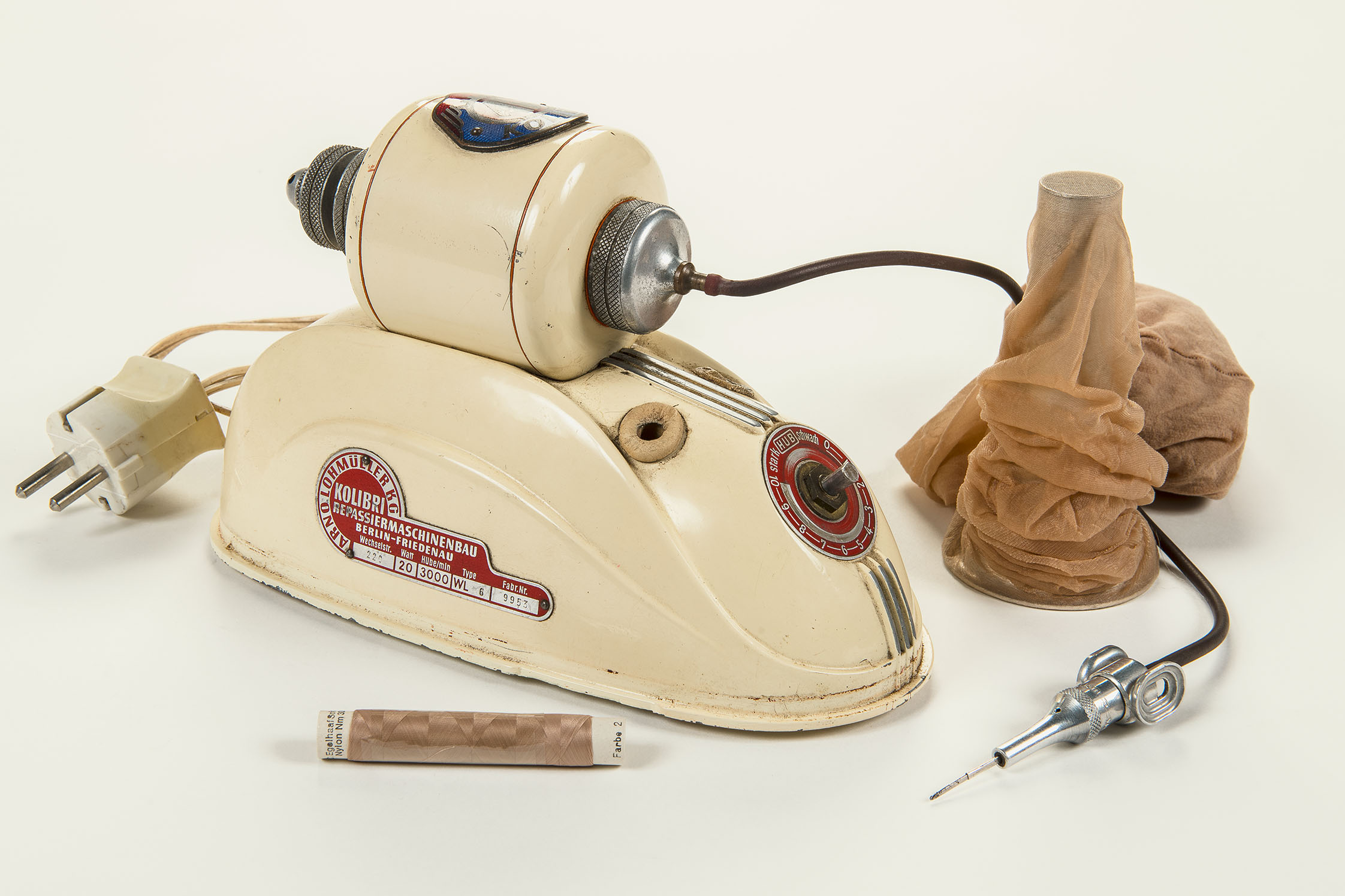 The image shows a cream-colored machine that vaguely resembles an iron. It has a wide base, a plug, a brown hose, and a silver-colored needle. Next to it is a stand holding a pair of beige-brown sheer pantyhose, and in front of it lies a spool of thread in the same color.