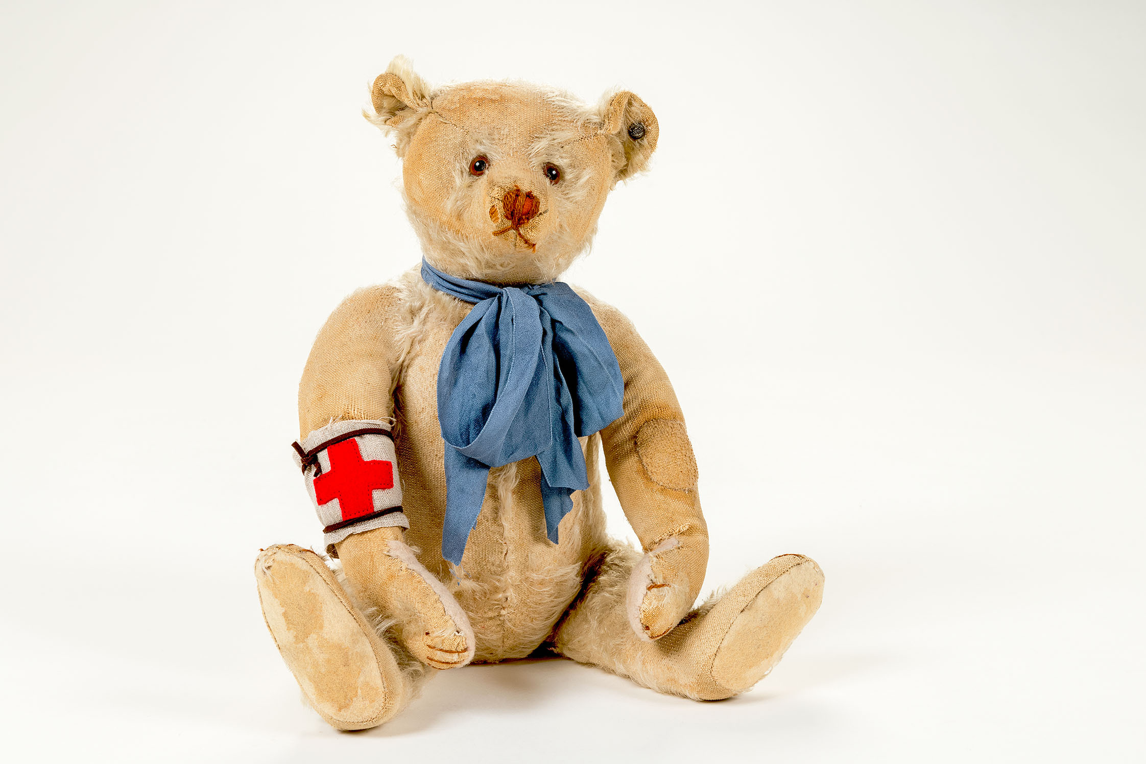 The picture shows a beige teddy bear with obvious signs of wear and tear. It is wearing a blue bow around its neck and a white armband with a red cross.
