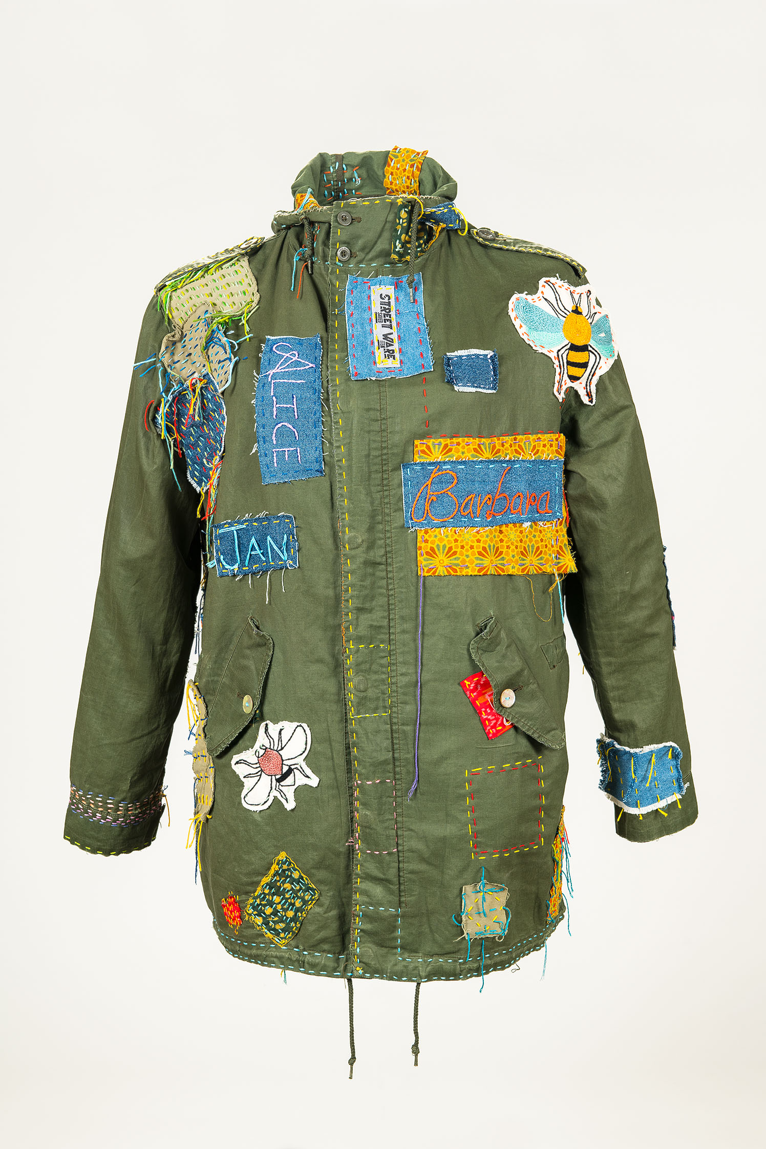 An olive-green outdoor jacket has been repaired with various eye-catching, colorful patches: Two iron-on patches featuring insect motifs stand out, as do large embroidered names on iron-on patches that identify members of the Berlin collective “STREETWARE saved item.”  