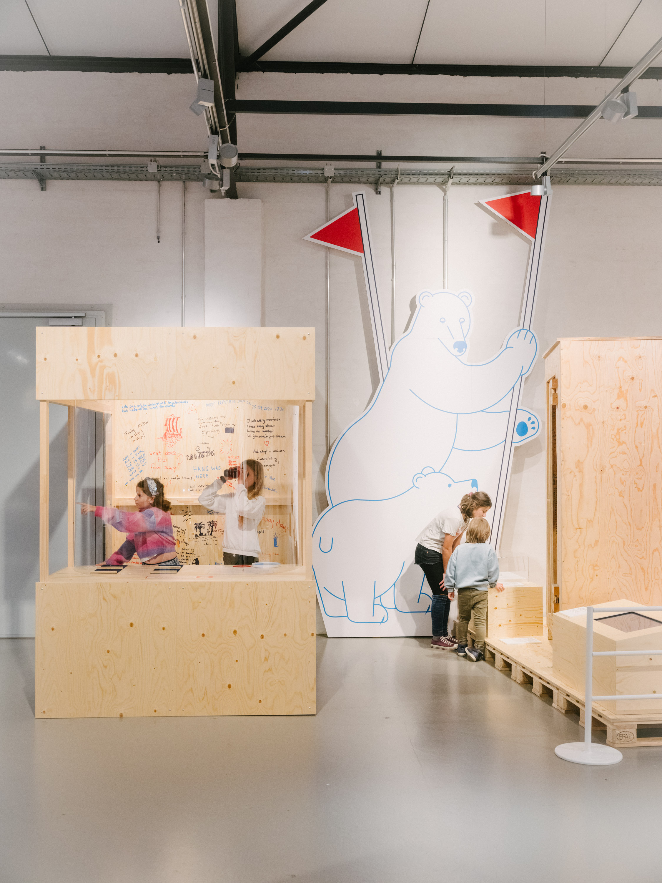 Two children are looking out of a roughly built wooden hut through a telescope. To the right of them are cartoon-like figures of two polar bears.