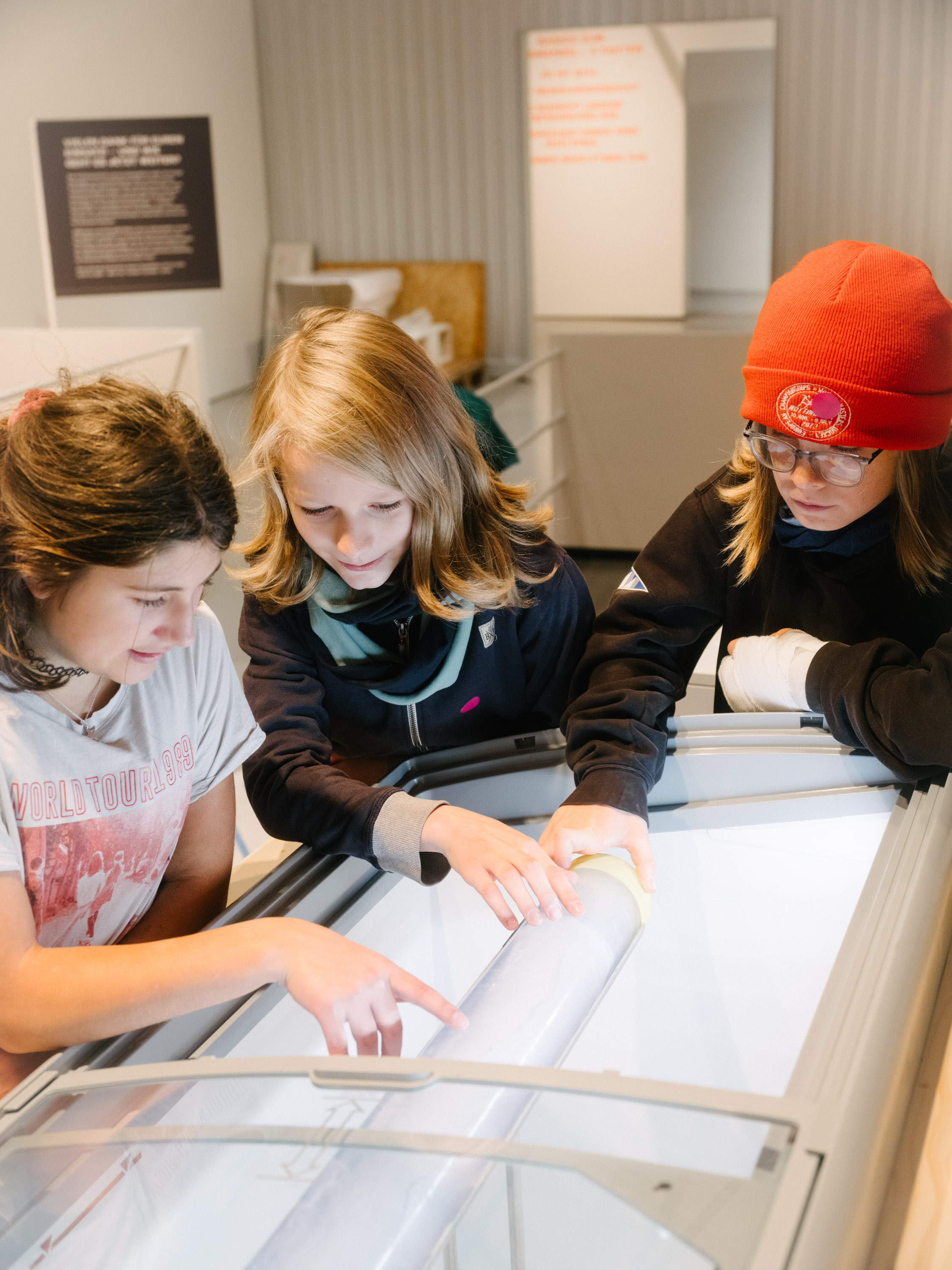 Drei Kinder betrachten und untersuchen einen zylinderförmigen Eisbohrkern in einer Ausstellungshalle.