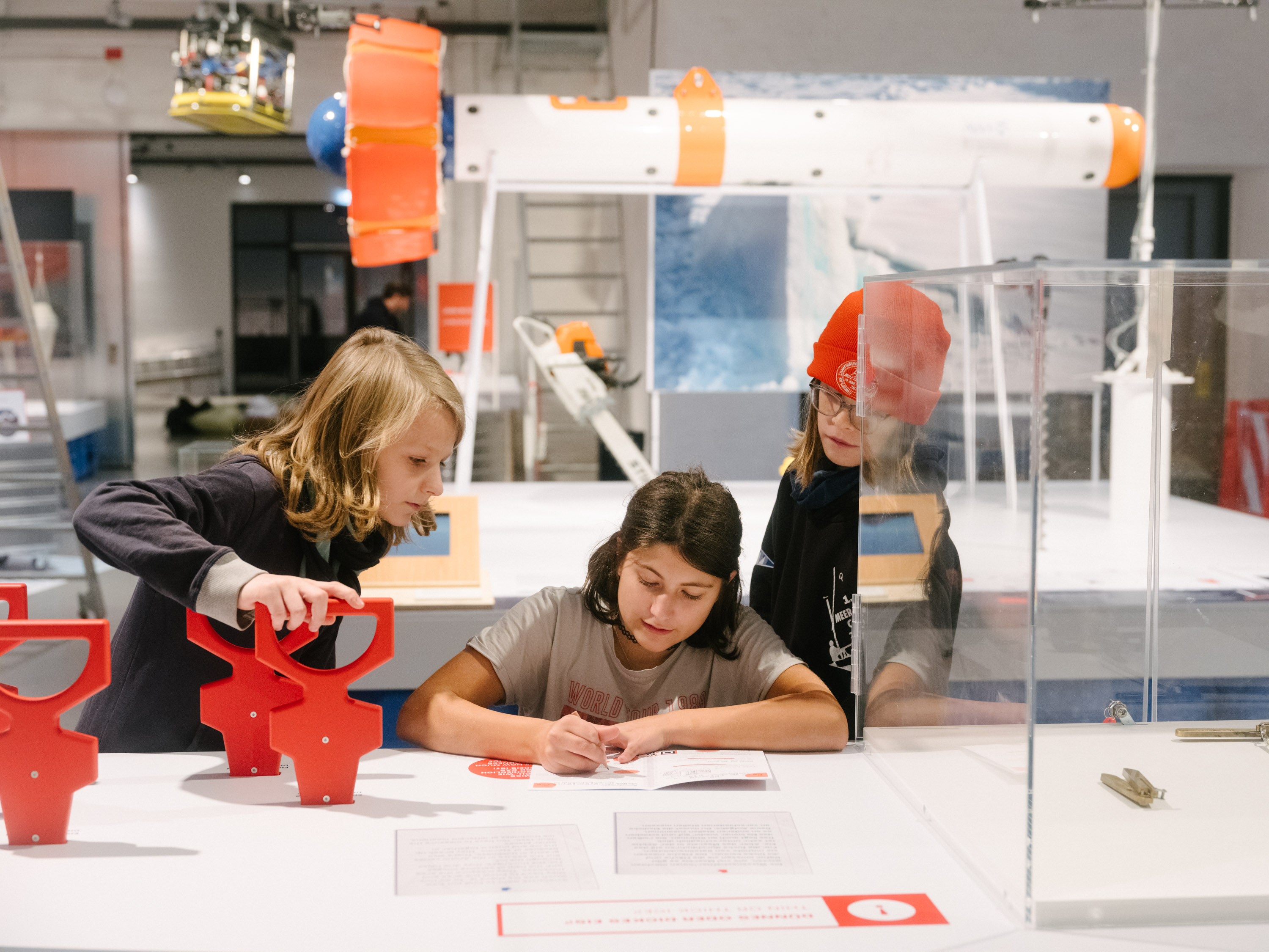 Three children are measuring the thickness of an "ice floe" at an exhibition table and recording their results in a research notebook.