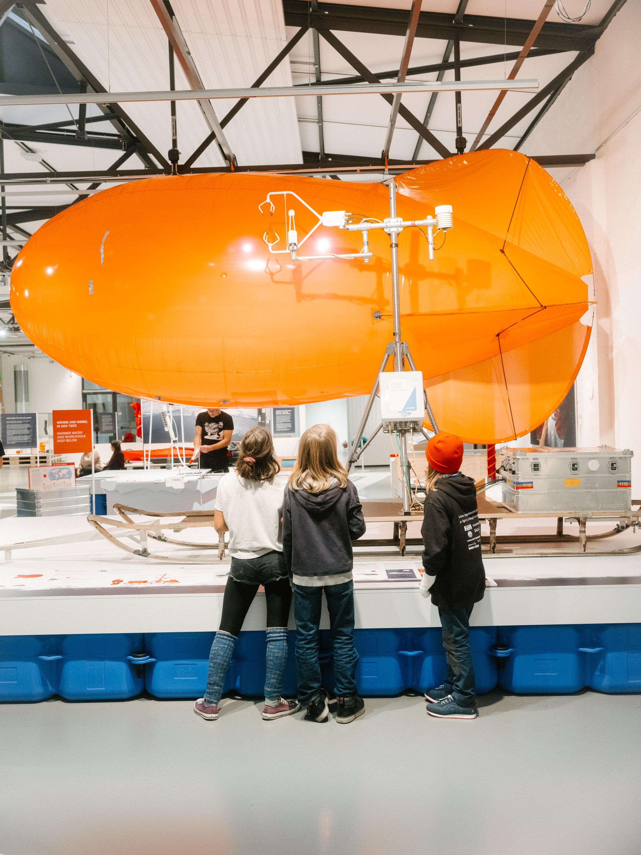 An orange tethered balloon hangs from the ceiling of an exhibition hall. Three children are standing in front of it, looking at it.
