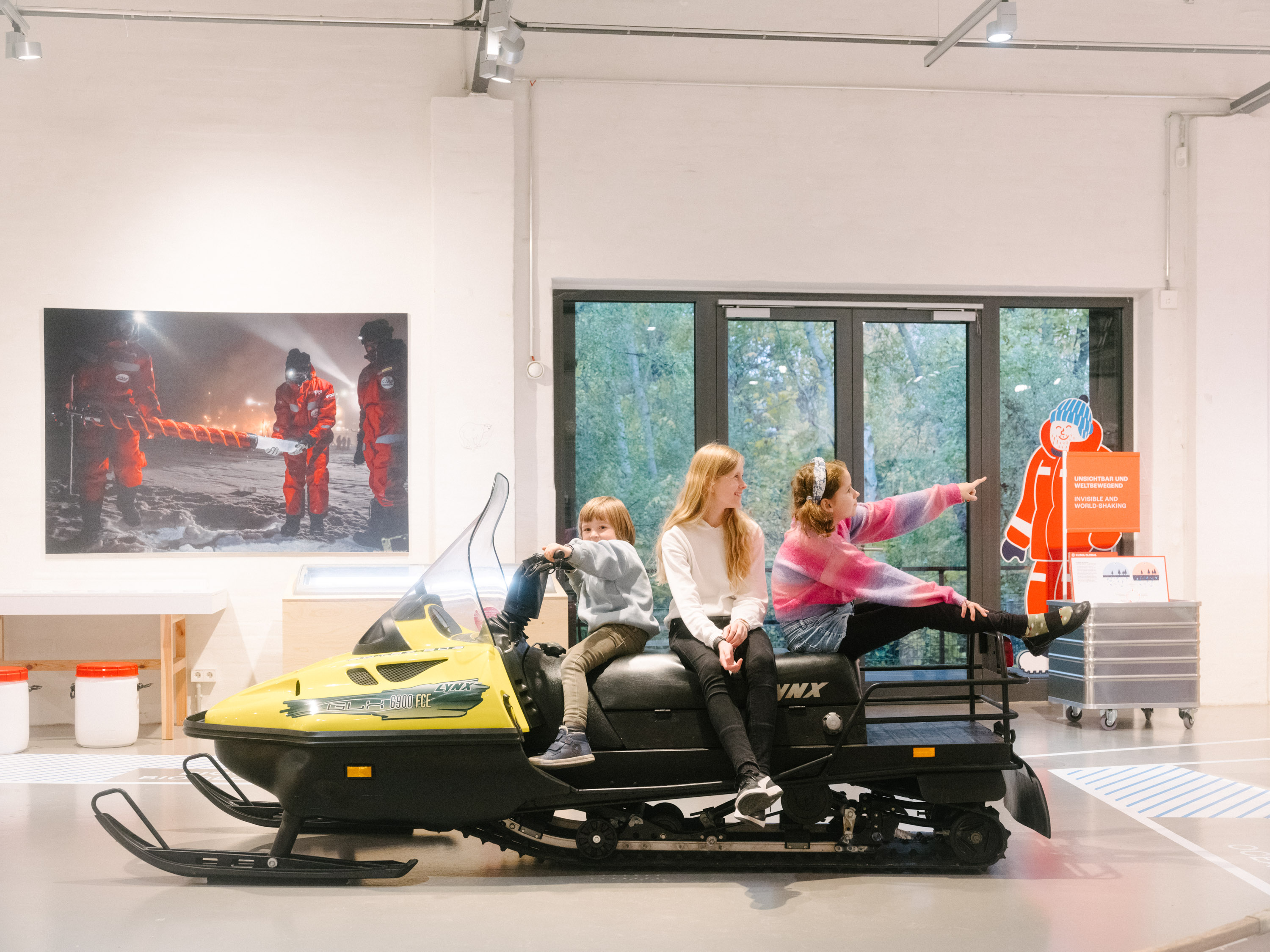 Three children are sitting on a black-and-yellow snowmobile in an exhibition hall.