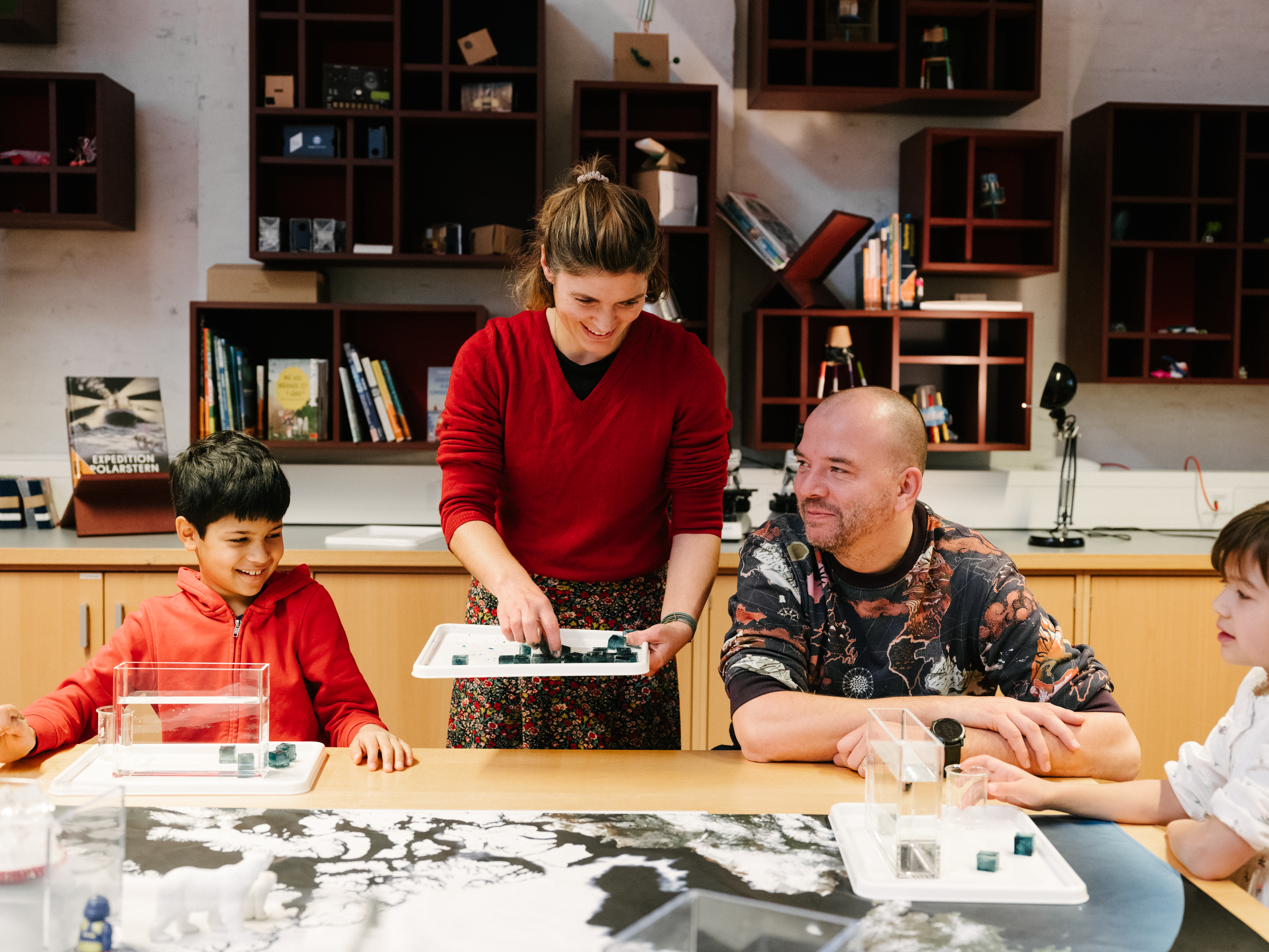 A family with two children is sitting and standing around a table, playing with ice cubes and water.