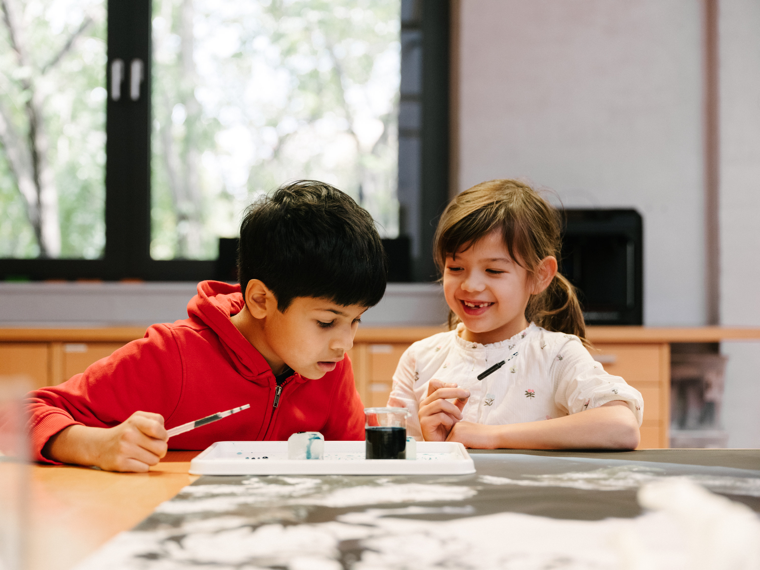 Two children are experimenting at a table with water, ice, paint, and pipettes.