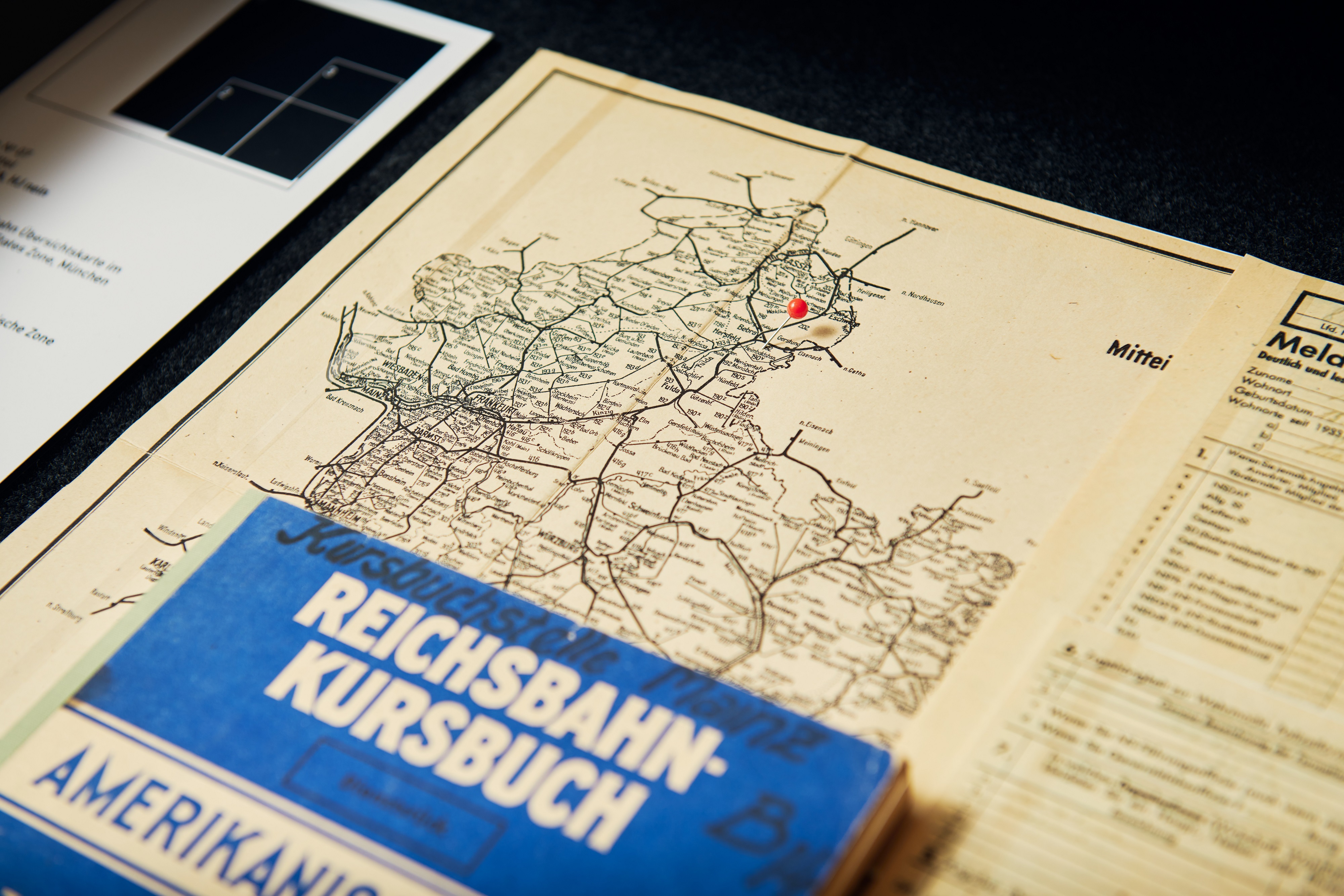 A map showing the railway network, with the cities of Frankfurt and Wiesbaden visible. On top of it lies a blue book labeled "Reichsbahn Kursbuch."