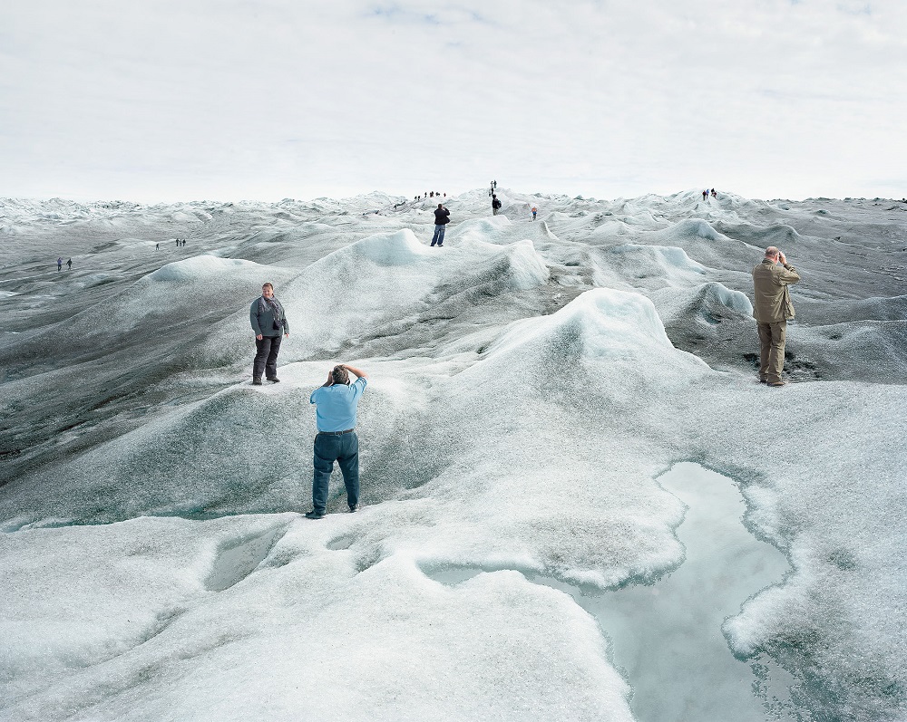 Eine Gruppe von Touristinnen und Touristen steht auf einer schmelzenden Eislandschaft und posiert an verschiedenen Punkten des Bildes für Fotos. An vielen Stellen der Eislandschaft hat sich schwarzer Ruß abgesetzt, das Eis ist dort sehr dunkel. Im Vordergrund ist eine große Pfütze aus Schmelzwasser.