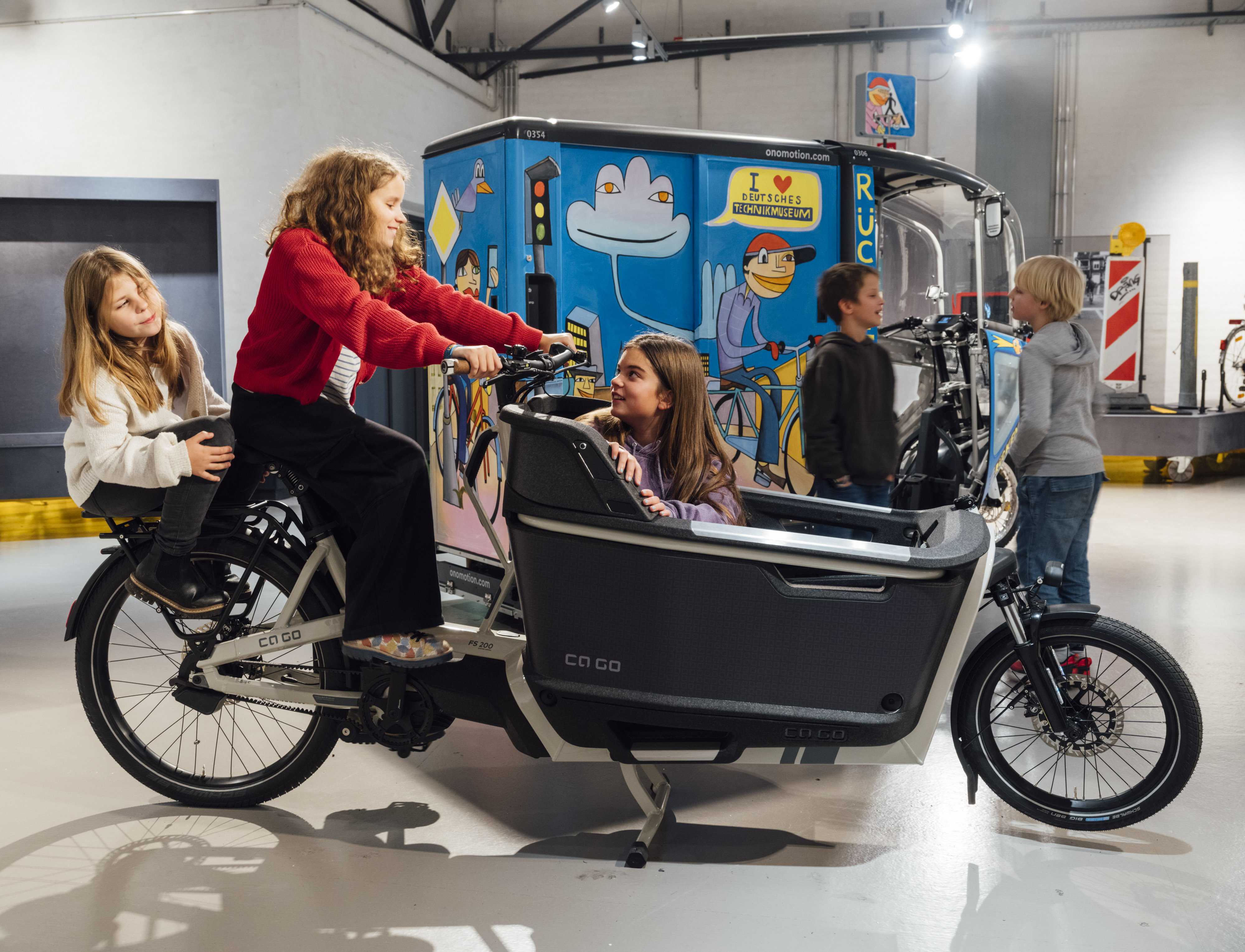 Blick in eine Ausstellung. Auf einem modernen Lastenrad sitzen zwei Mädchen, ein weiteres Mädchen sitzt in der Ladefläche. Im Hintergrund unterhalten sich zwei Jungen vor einem bunten großen E-Lastenrad.