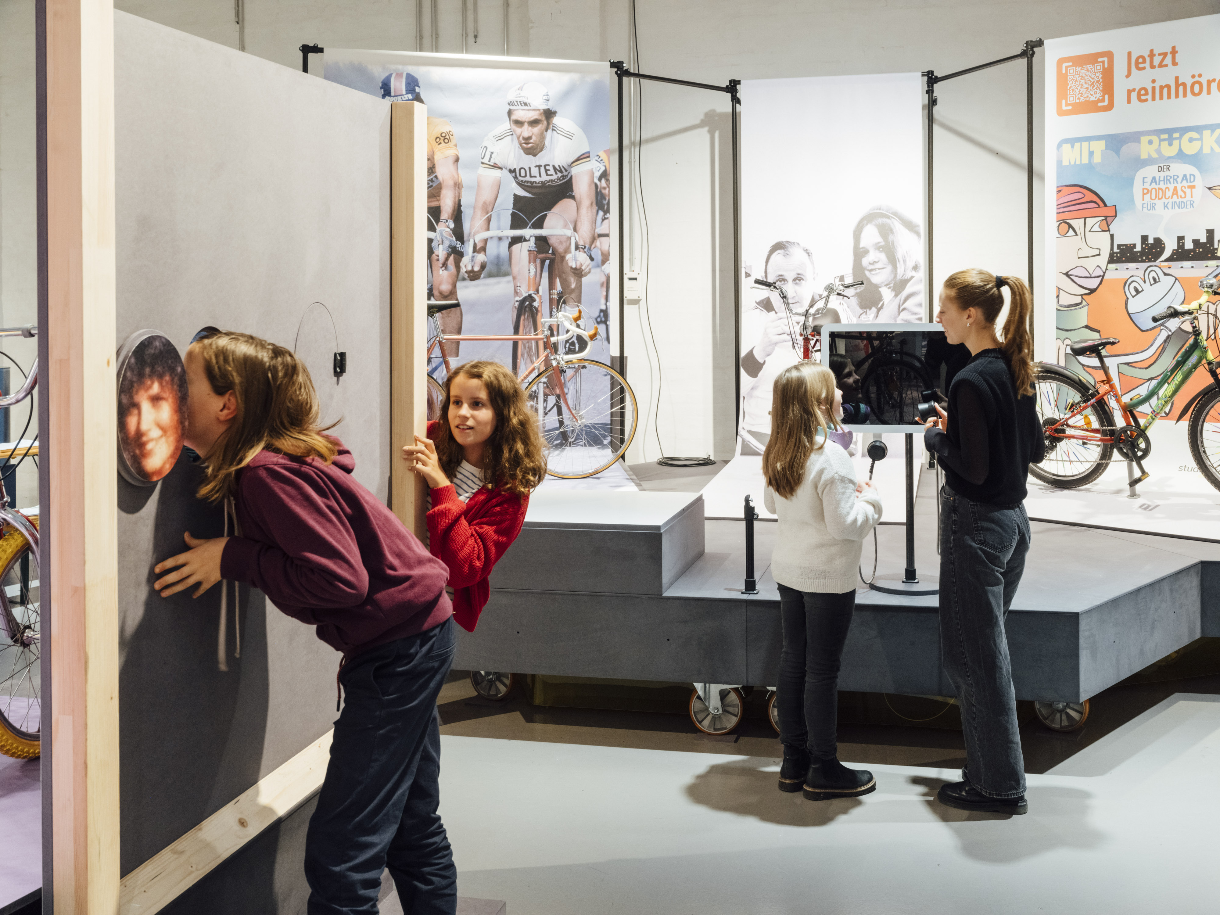 Mehrere Menschen in einem Ausstellungsraum. Im Vordergrund zwei junge Mädchen, die sich mit einer Holzwand mit Löchern darin beschäftigen, im Hintergrund eine junge Frau und ein kleines Mädchen, die sich vor einer Ausstellungseinheit mit Fahrrädern und Abbildungen von Fahrradfahrern unterhalten.