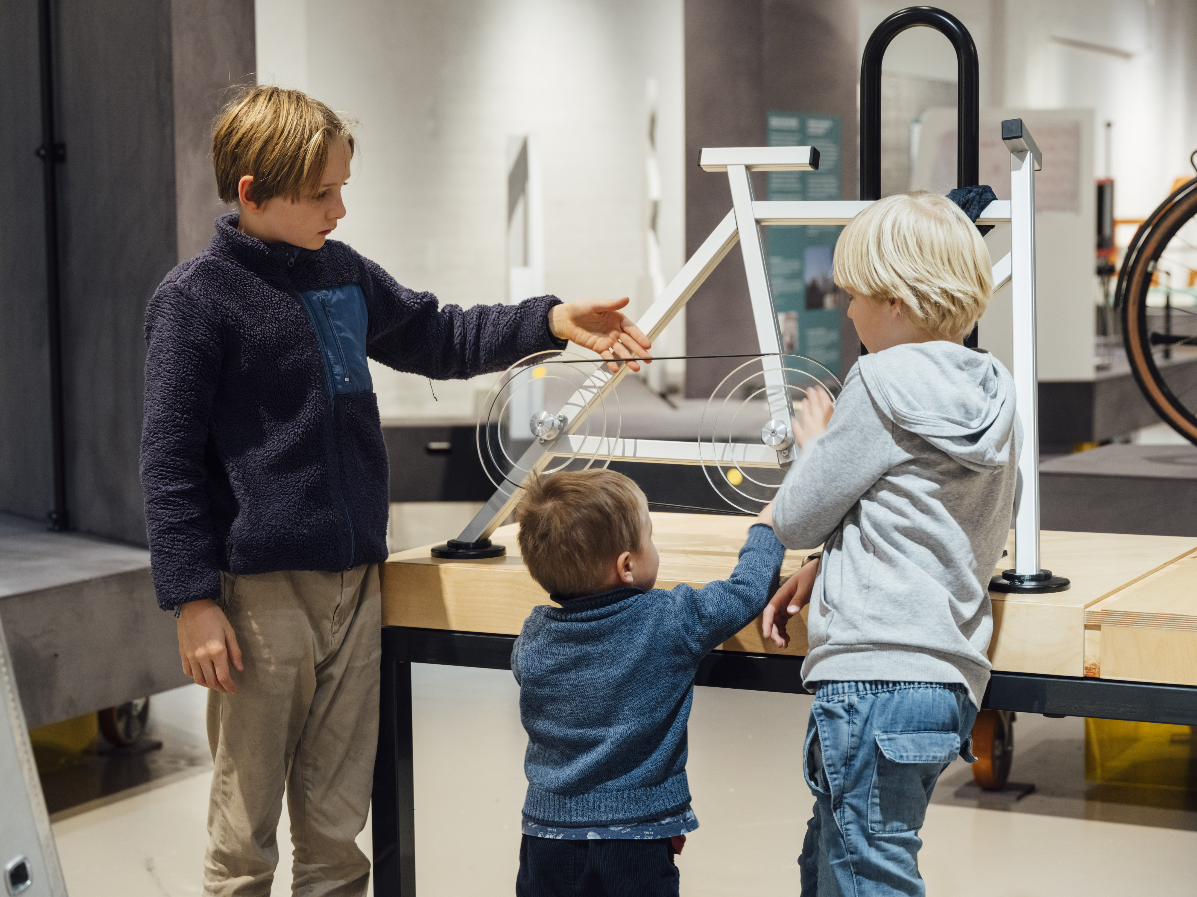 Drei Kinder stehen in einem Ausstellungsraum vor einem schematisch vereinfachten Nachbau eines Fahrrads. Ein Junge berührt die dort dargestellte Fahrradkette.