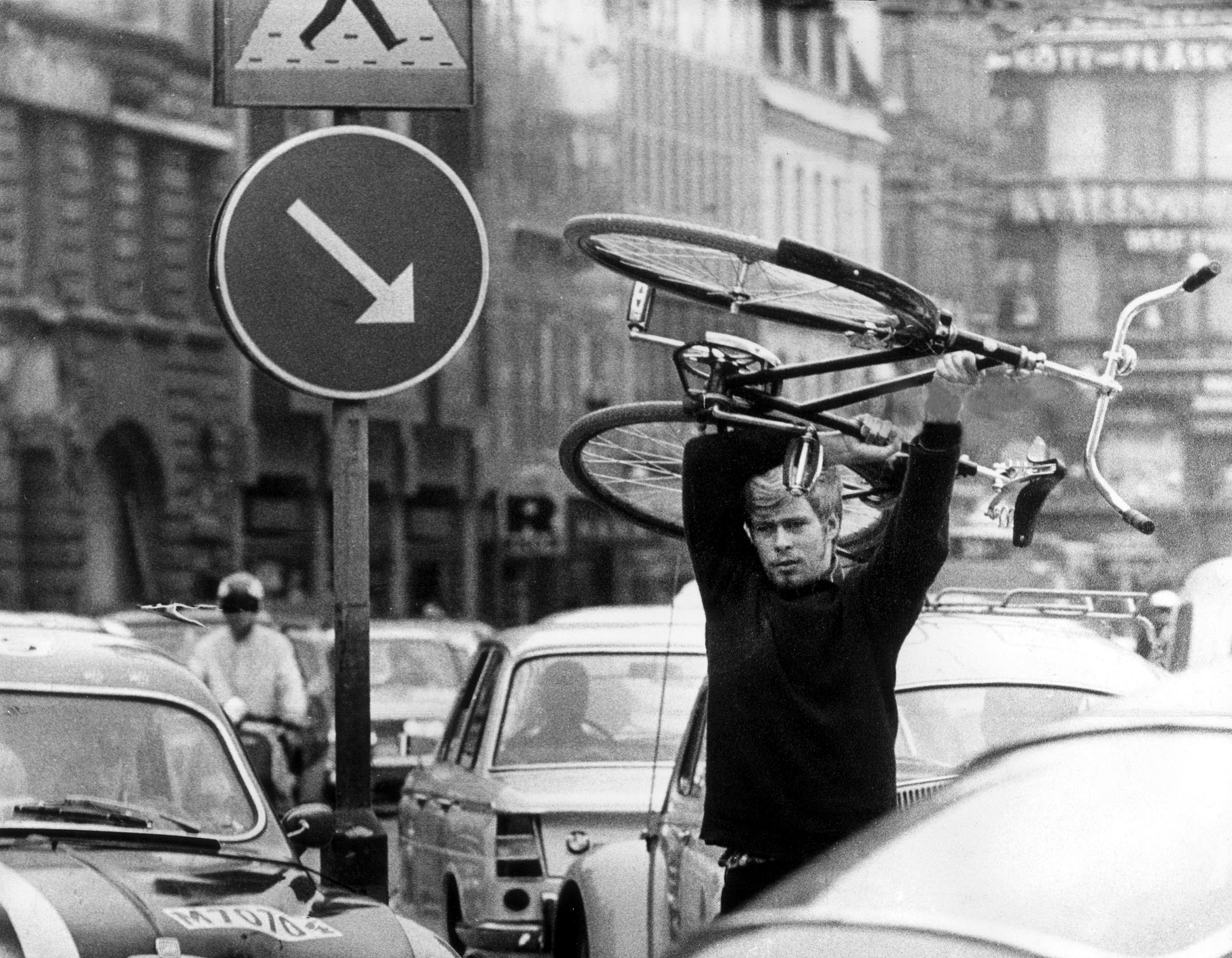 Ein Mann trägt ein Fahrrad über seinem Kopf. Er läuft zwischen im Stau stehenden Autos hindurch. Das Schwarz-Weiß-Foto wurde am 3.09.1967 in Malmö aufgenommen.  An diesem Tag stellte Schweden von Links- auf Rechtsverkehr um. In Teilen des Landes führte das zunächst zu Problemen und Staus.