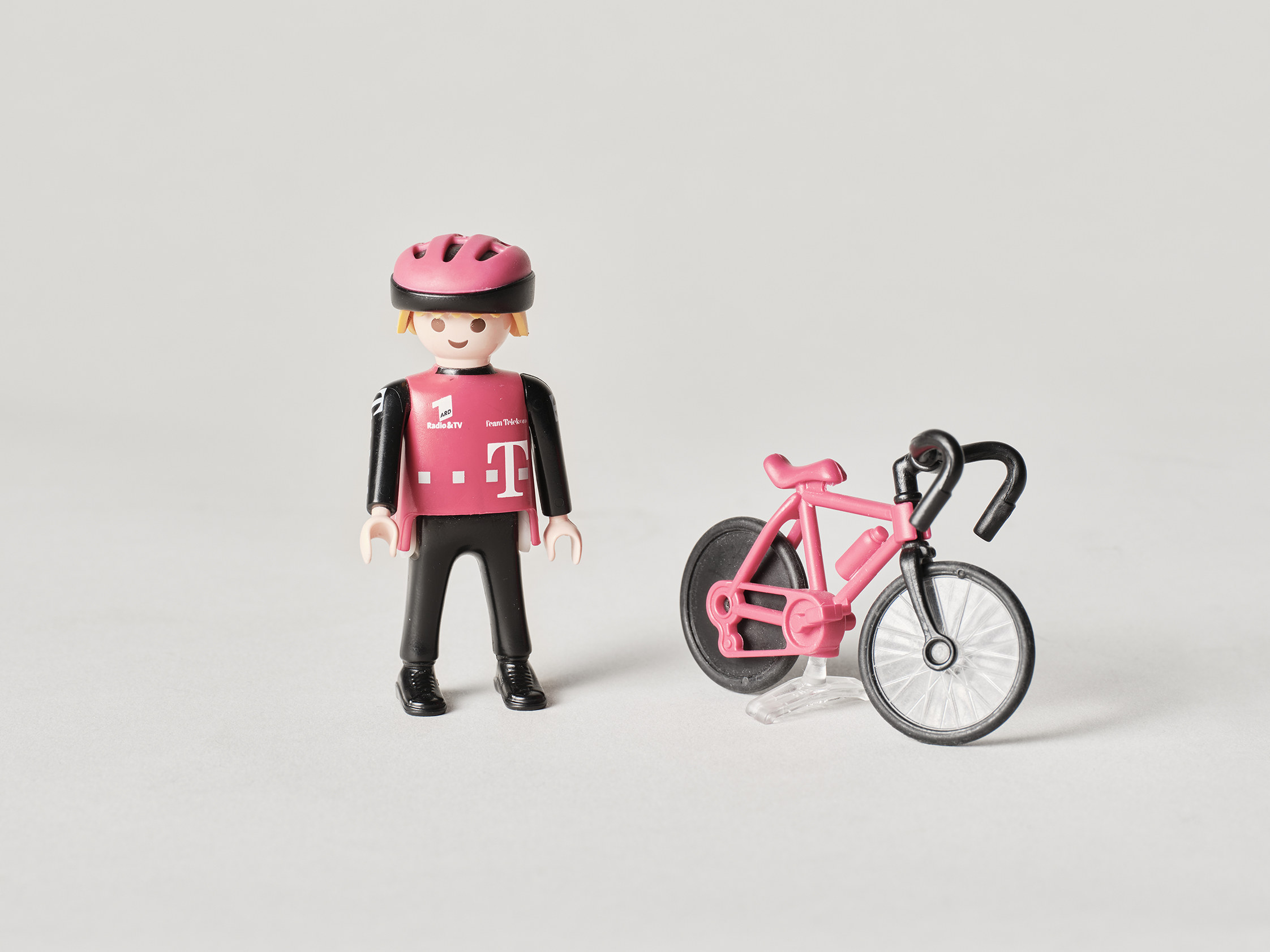 Das Foto zeigt eine Spielzeugfigur aus Kunststoff vom Hersteller Playmobil. Es ist ein Radfahrer mit einem Trikot und einer Mütze des Radsportteams von Telekom. Trikot und Mütze haben die Team Telekom Farbe Magenta. Neben der Figur ist ein Rennrad zu sehen. Der Rahmen des Fahrrads ist ebenfalls magentafarben.