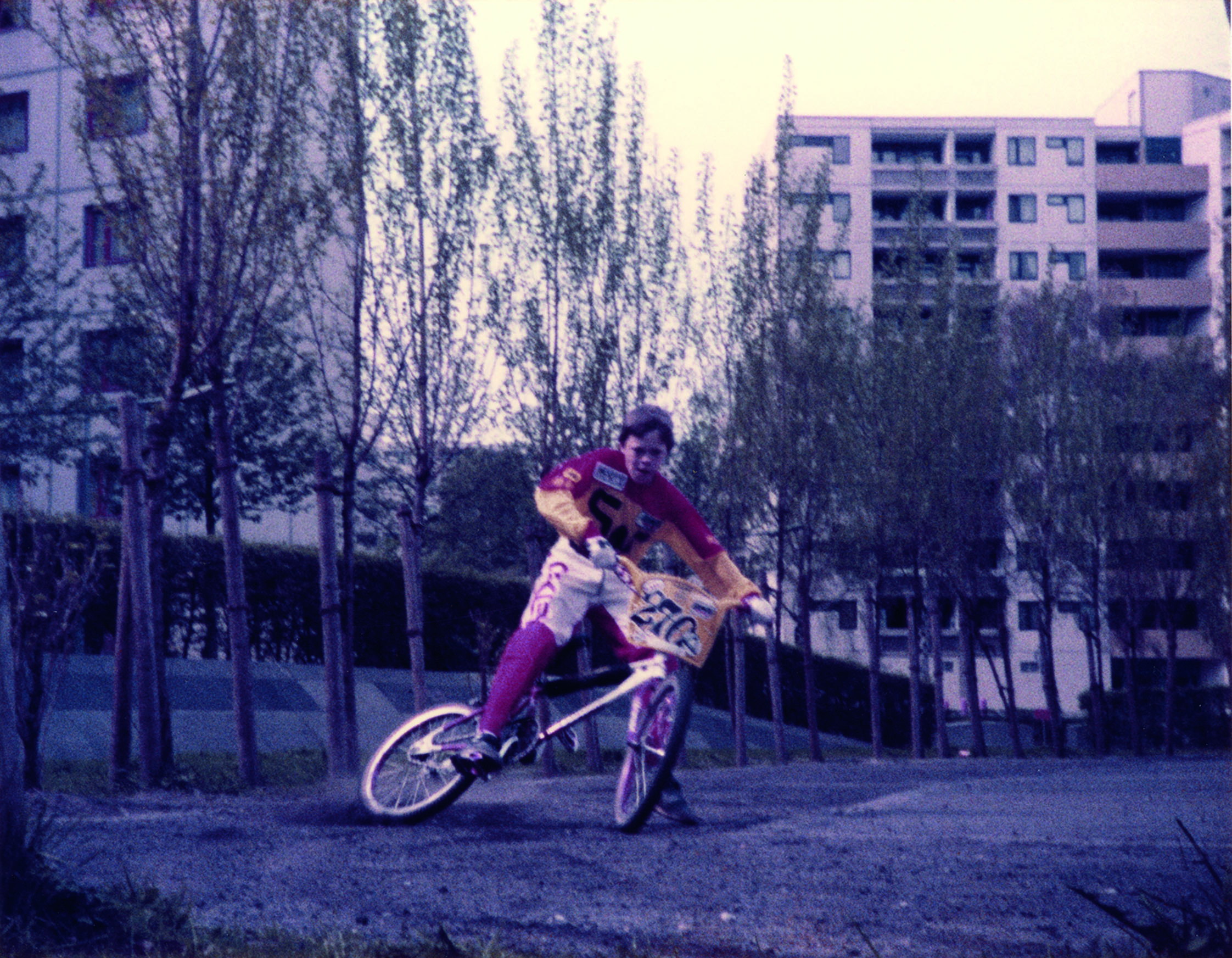Ein Kind fährt vor dem Hintergrund von Hochhäusern auf einem BMX-Fahrrad. Er bremst gerade und driftet zur Seite. Das Kind trägt eine Hose und ein Shirt aus dem BMX-Sport, aber keinen Helm, das BMX-Rad hat eine Start-Nummer für Sportwettbewerbe am Lenker.