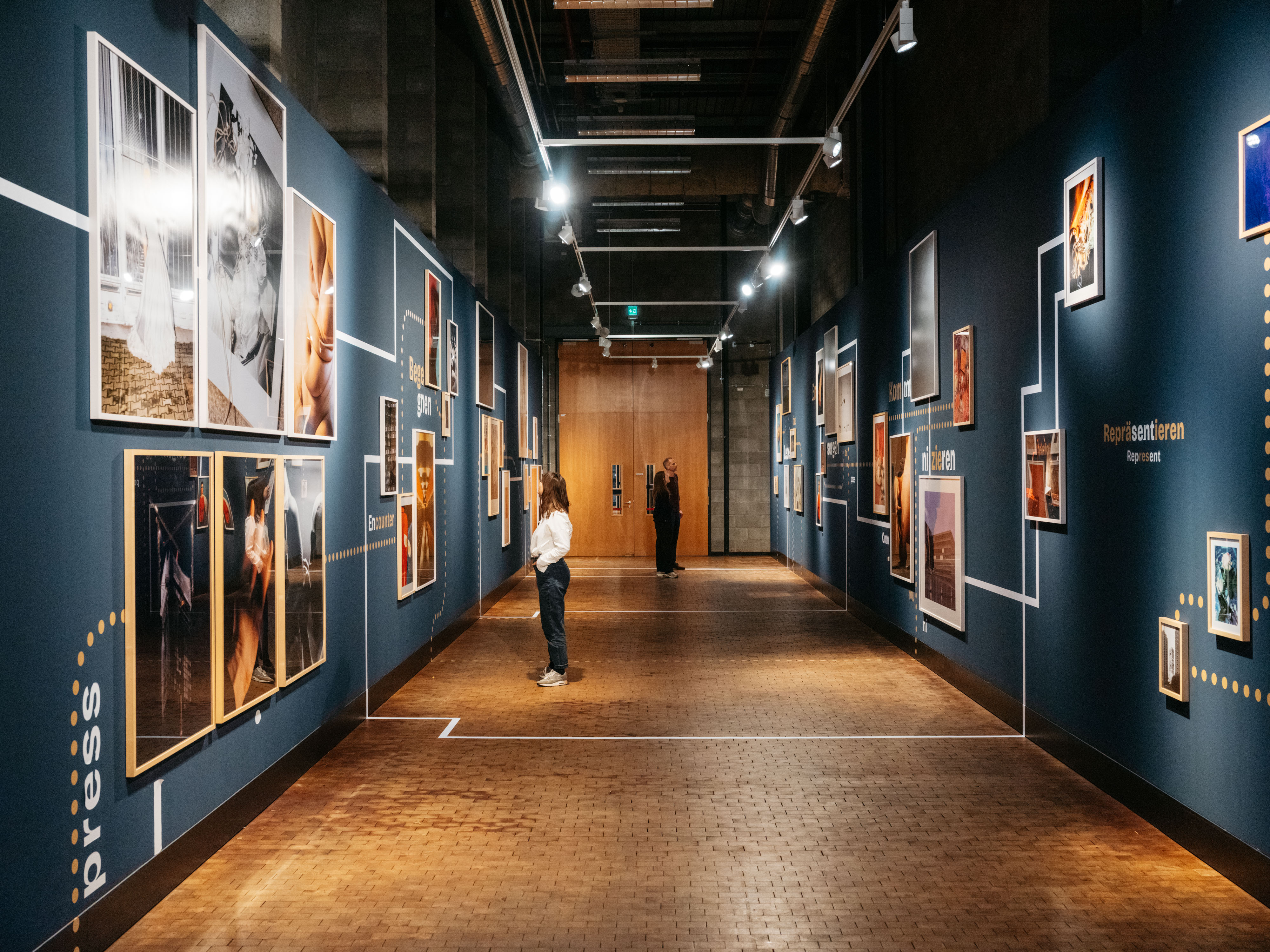 Blick in einen langen Ausstellungsraum mit dunkelblauen Wänden. Es hängen gerahmte Fotografien an den Wänden. Zwischen den Fotos sind Verbindungslinien und gepunktete Verbindungslinien erkennbar. Einzelne Personen stehen vor den Bildern und betrachten diese.